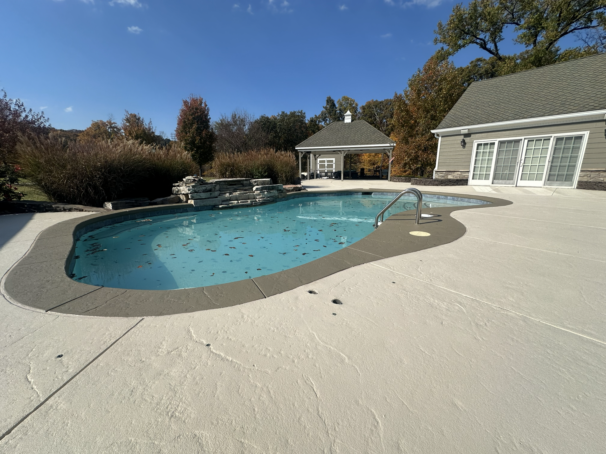A backyard with a kidney-shaped swimming pool surrounded by a white concrete deck. There are fallen leaves floating on the water. In the background, trees with fall foliage, a small stone waterfall, a gazebo, and a house with sliding glass doors are visible under a clear blue sky.