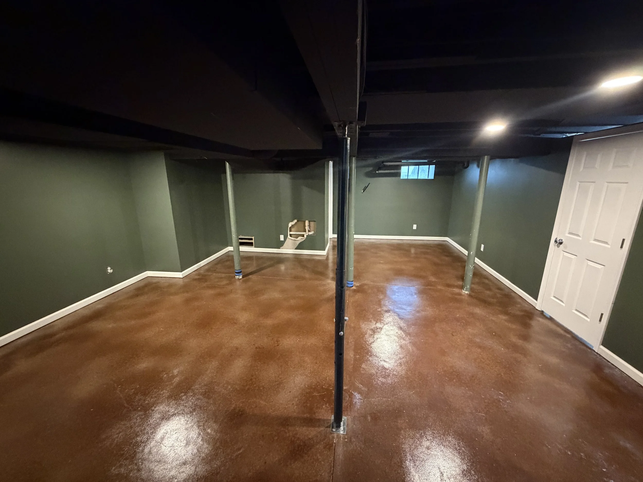 Empty basement room with green walls, a small window, wooden floors, a white door, and support poles.