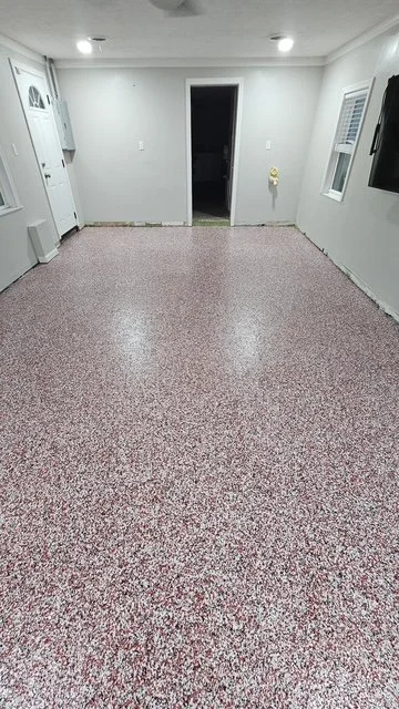 Empty room with white walls, a doorway leading outside, a window, electrical outlets, and speckled red and white flooring.