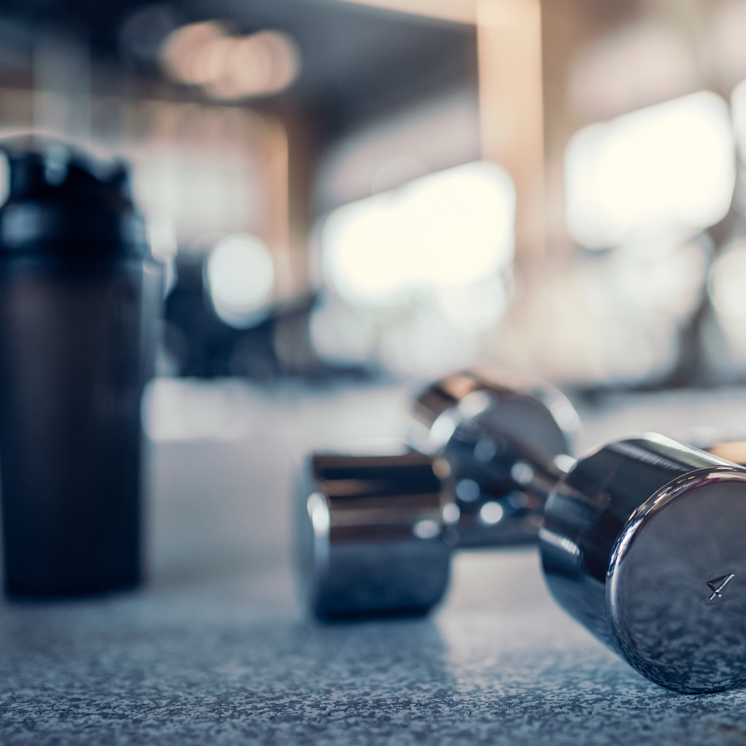 Close-up of a dumbbell and a shaker bottle on a gym bench with gym equipment blurred in the background.