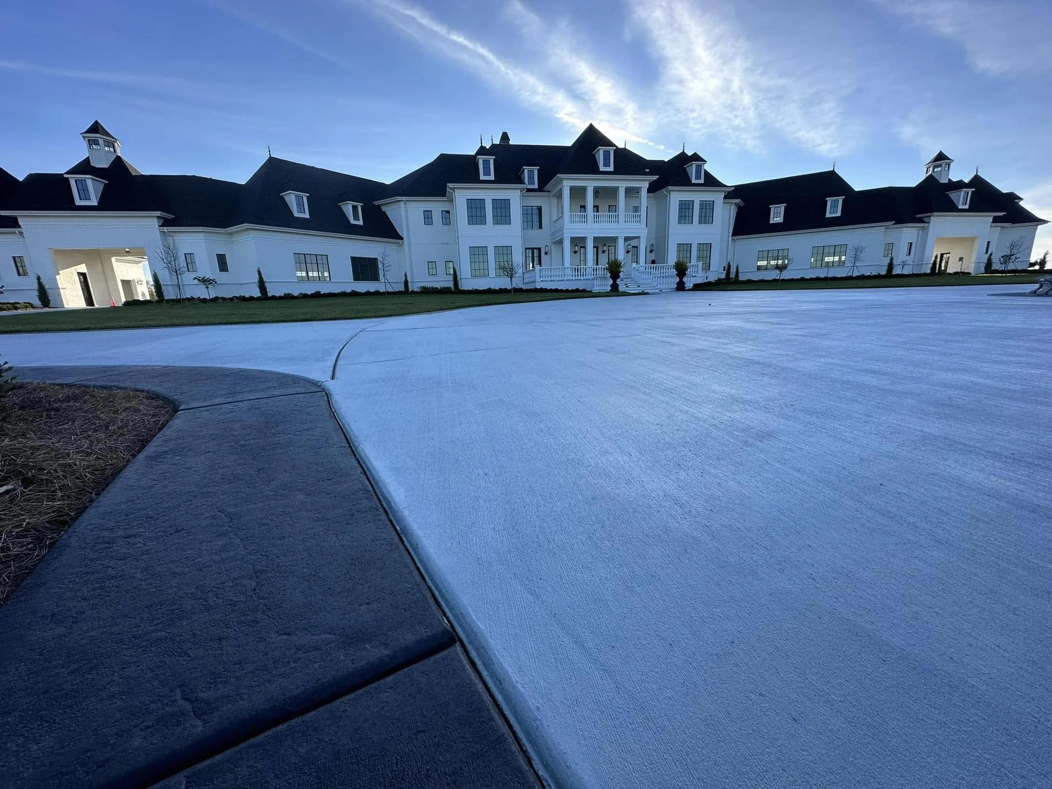 Large white mansion with black roof, multiple windows, and front porch, surrounded by a vast paved driveway and green lawn under a blue sky.