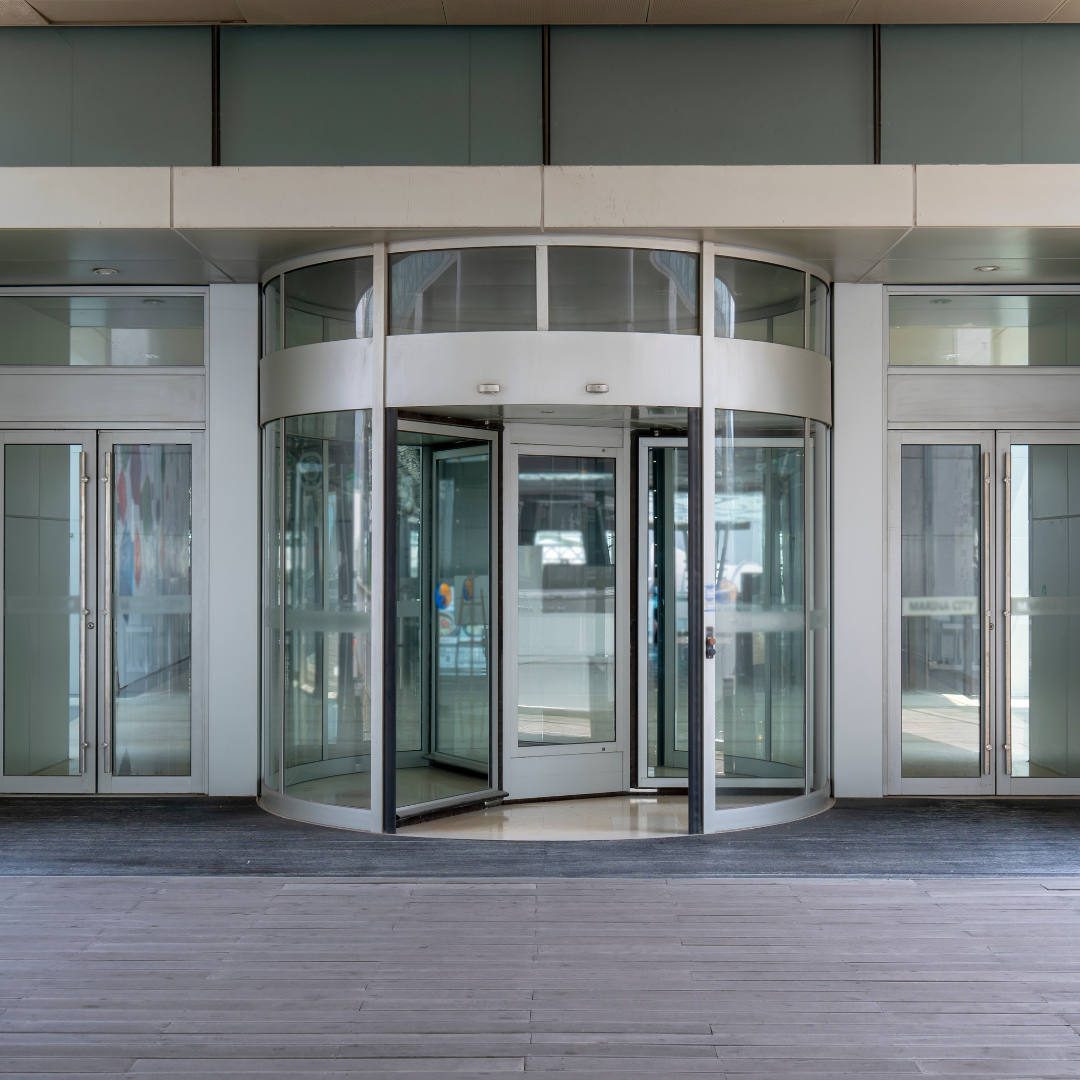 Front entrance of a modern building with glass revolving doors and side doors, with a wooden floor outside.