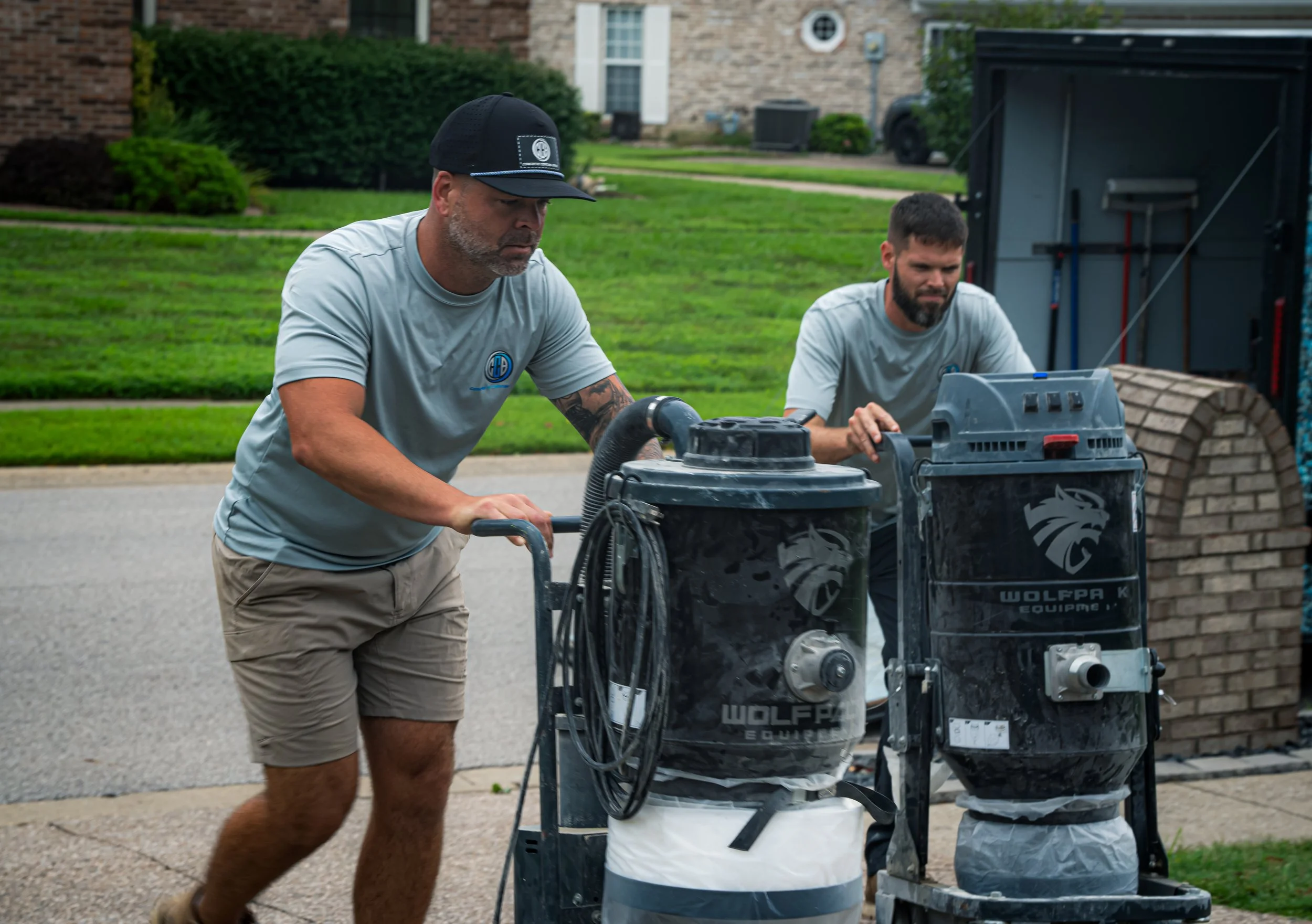 Two men working outside with large equipment on a sidewalk, one pushing a cart with machinery, and the other operating controls, in front of a house with a brick wall and green lawn.