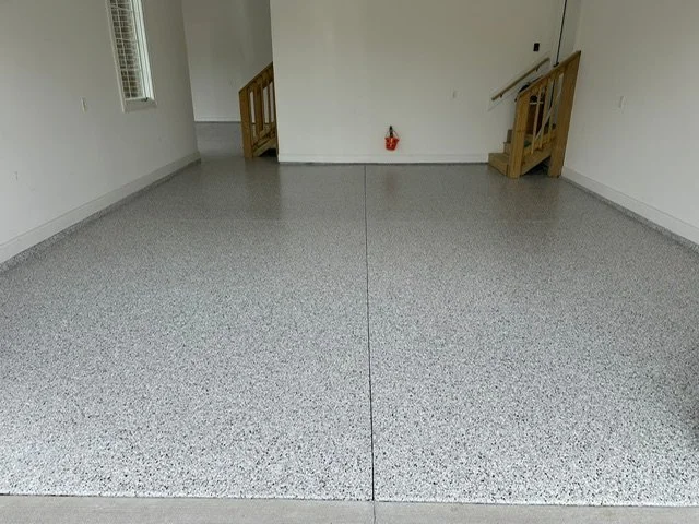 Empty room with speckled gray epoxy flooring, white walls, a small window, staircase with wooden railing, and a red fire extinguisher on the wall.