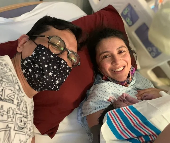 A woman in a hospital bed holding a newborn baby, smiling, with a man beside her wearing glasses and a star-patterned face mask, taking a selfie.