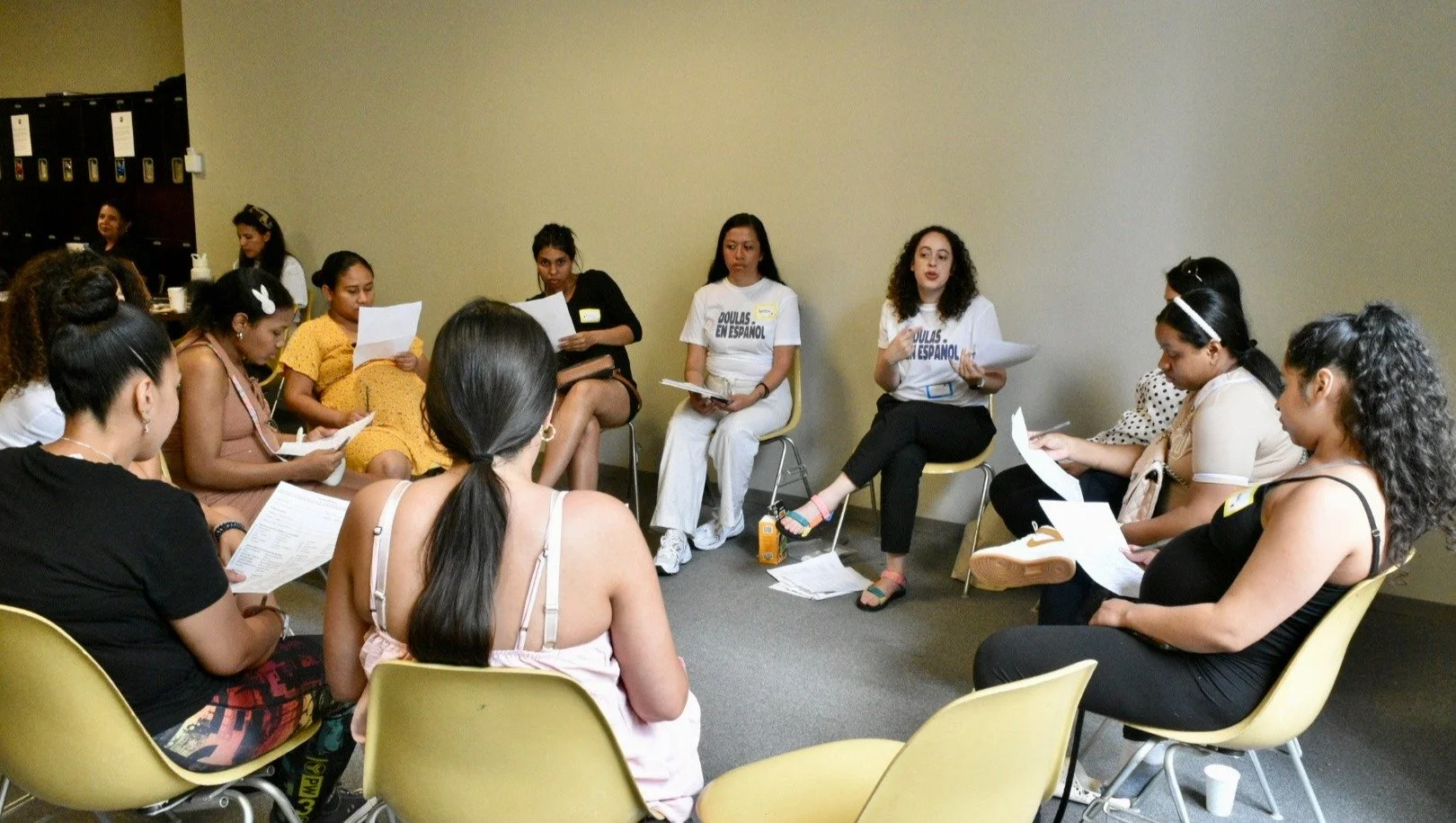 A group of women sitting in a circle, engaged in a discussion or meeting, some holding papers, with a plain wall in the background.