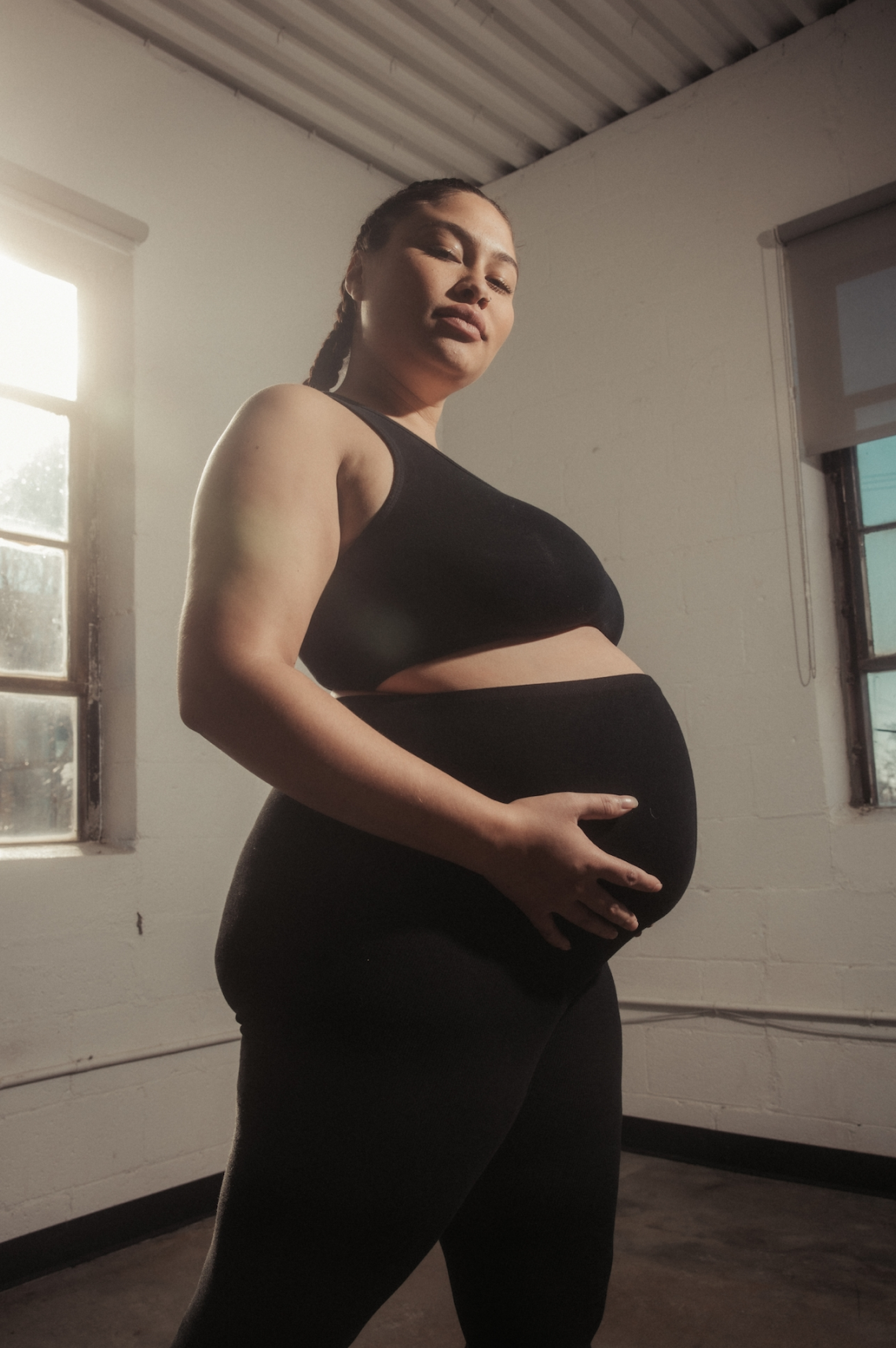Pregnant woman in black workout clothes standing indoors near a window, holding her belly with one hand, with sunlight coming through the window.