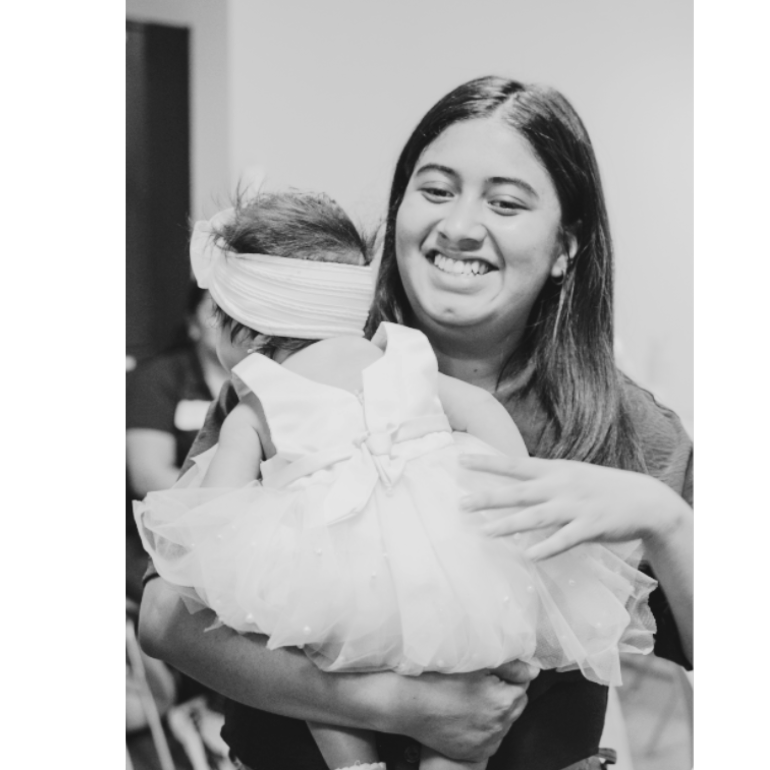 A woman smiling while holding a young child in a dress, with the child's face partially covered by a headband, in an indoor setting.