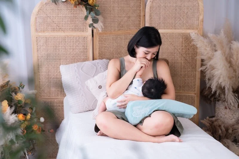 A woman breastfeeding a baby while sitting on a bed in a cozy room with floral decorations and naturalrattan headboard.