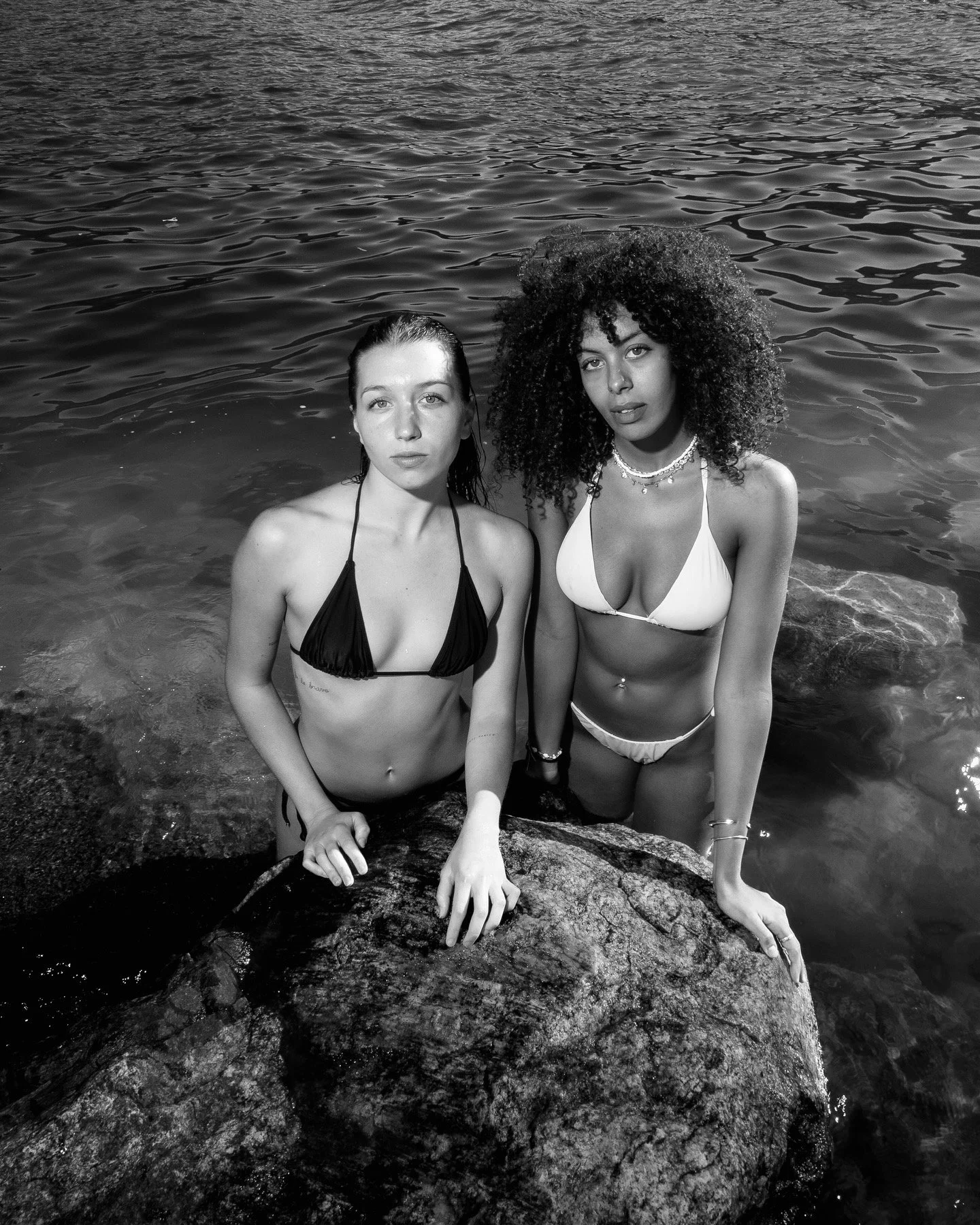 Two women in bikinis standing on rocks in water, posing for the camera, black and white photo.