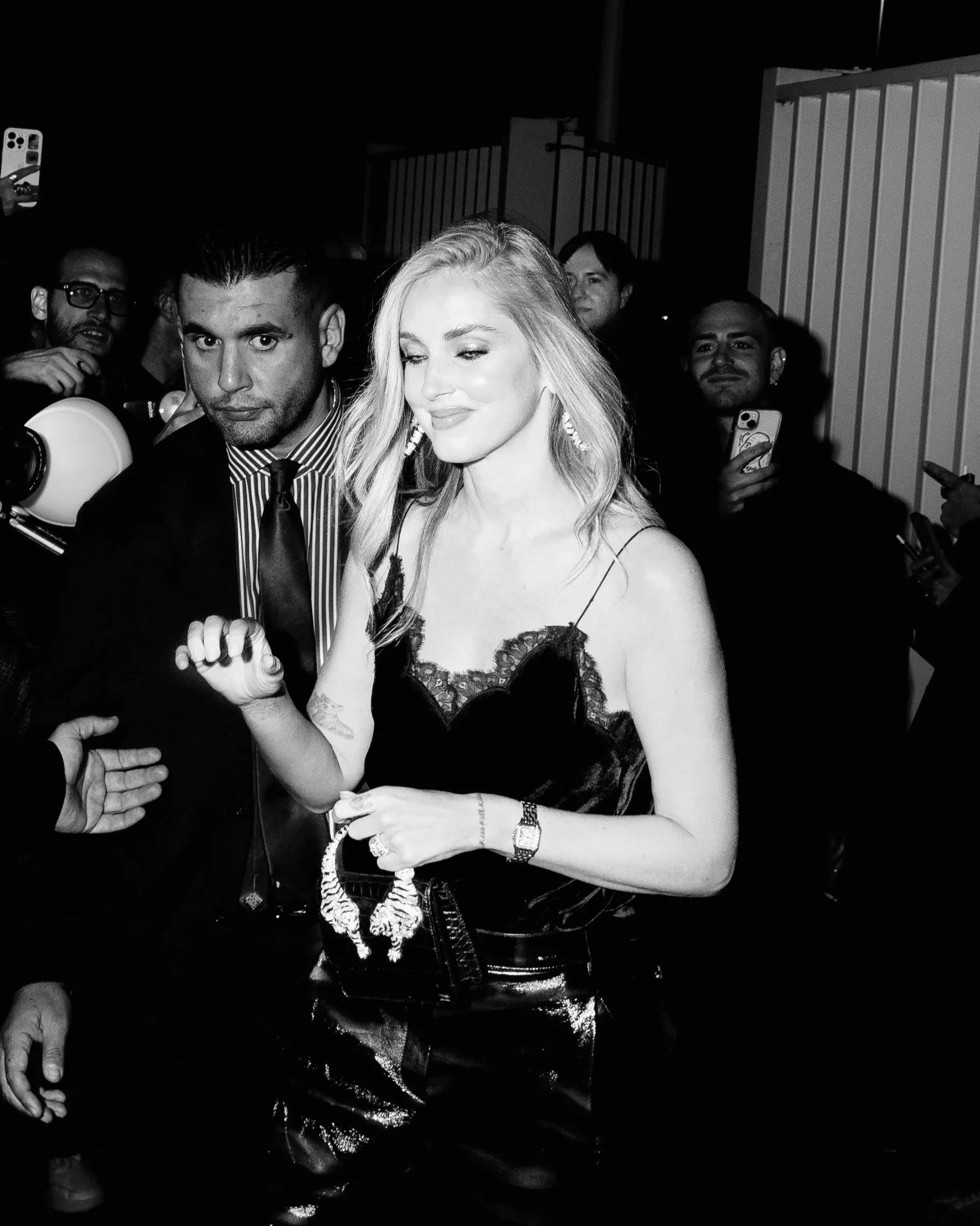 Chiara Ferragni, in a black lace top and shiny pants amid a crowd, smiling and holding a necklace, with people around taking photos at night.