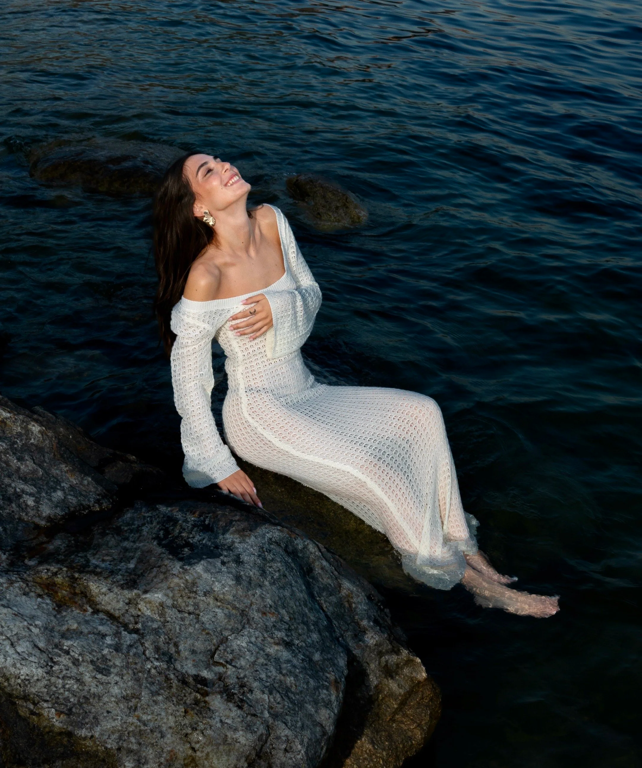 A woman in a white crocheted dress sitting on a large rock by the water, smiling with her head tilted back.