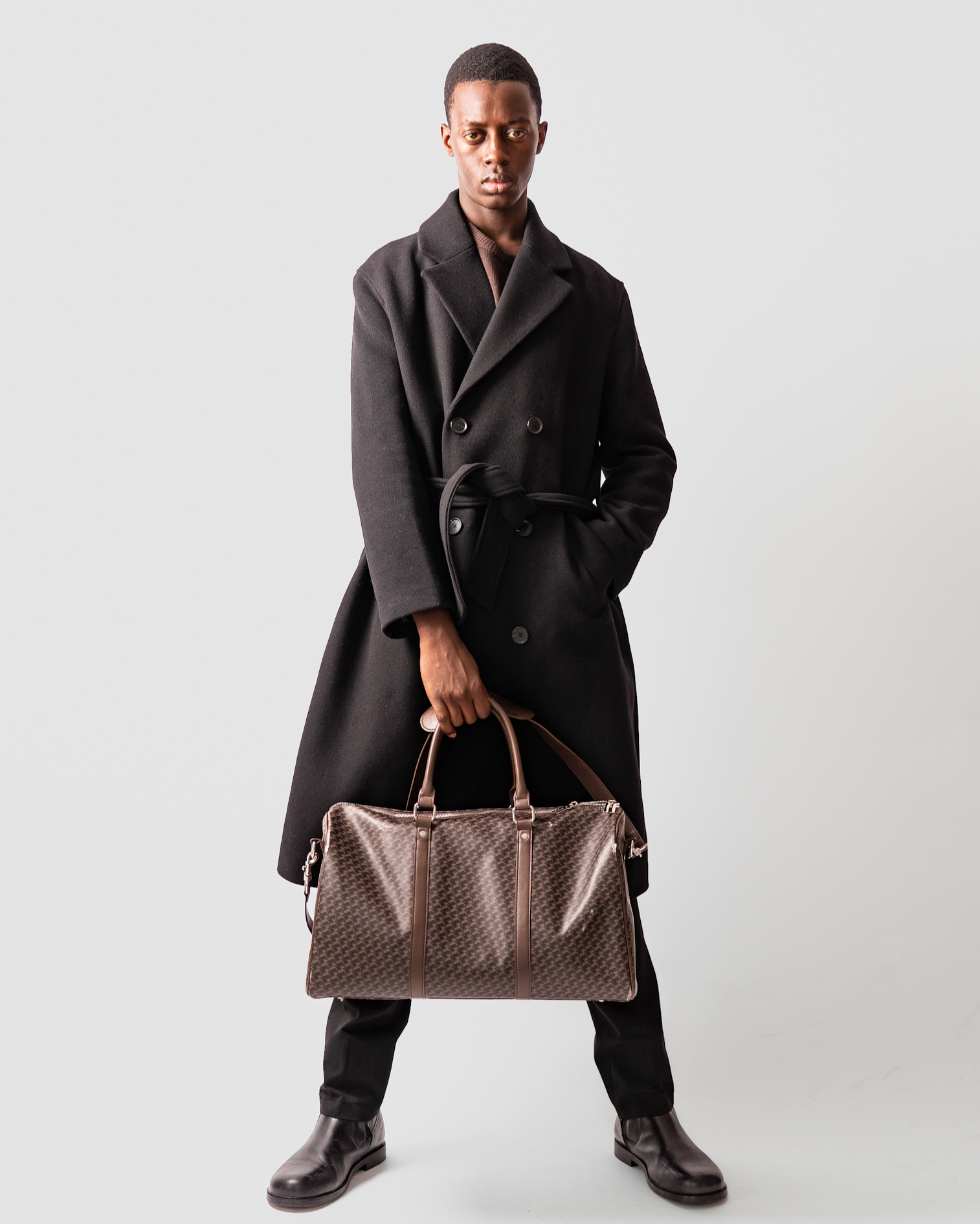 A young man standing against a plain light gray background, wearing a black coat with a belt, black pants, and black shoes. He is holding a brown patterned duffel bag in his right hand and has a serious expression.