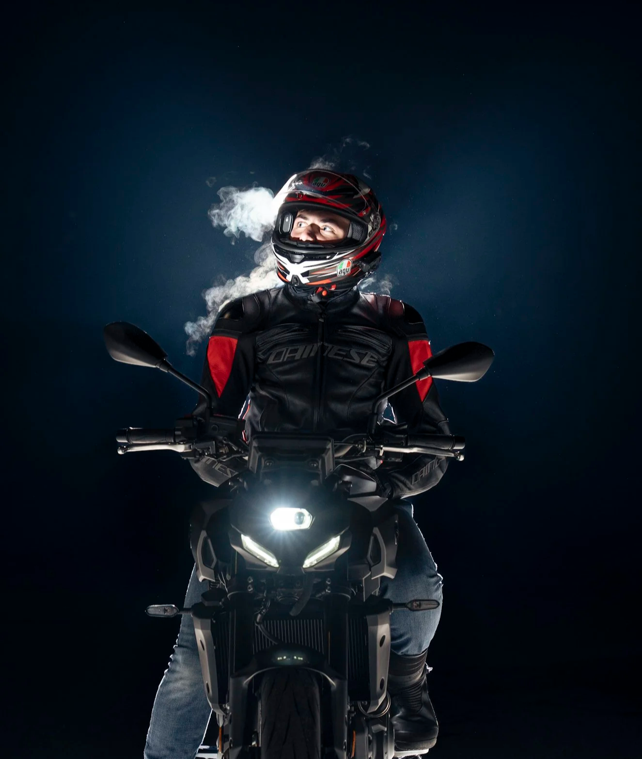 A person wearing a motorcycle helmet and jacket, sitting on a black motorcycle with headlights on, against a dark background, with visible vapor or steam near their face.