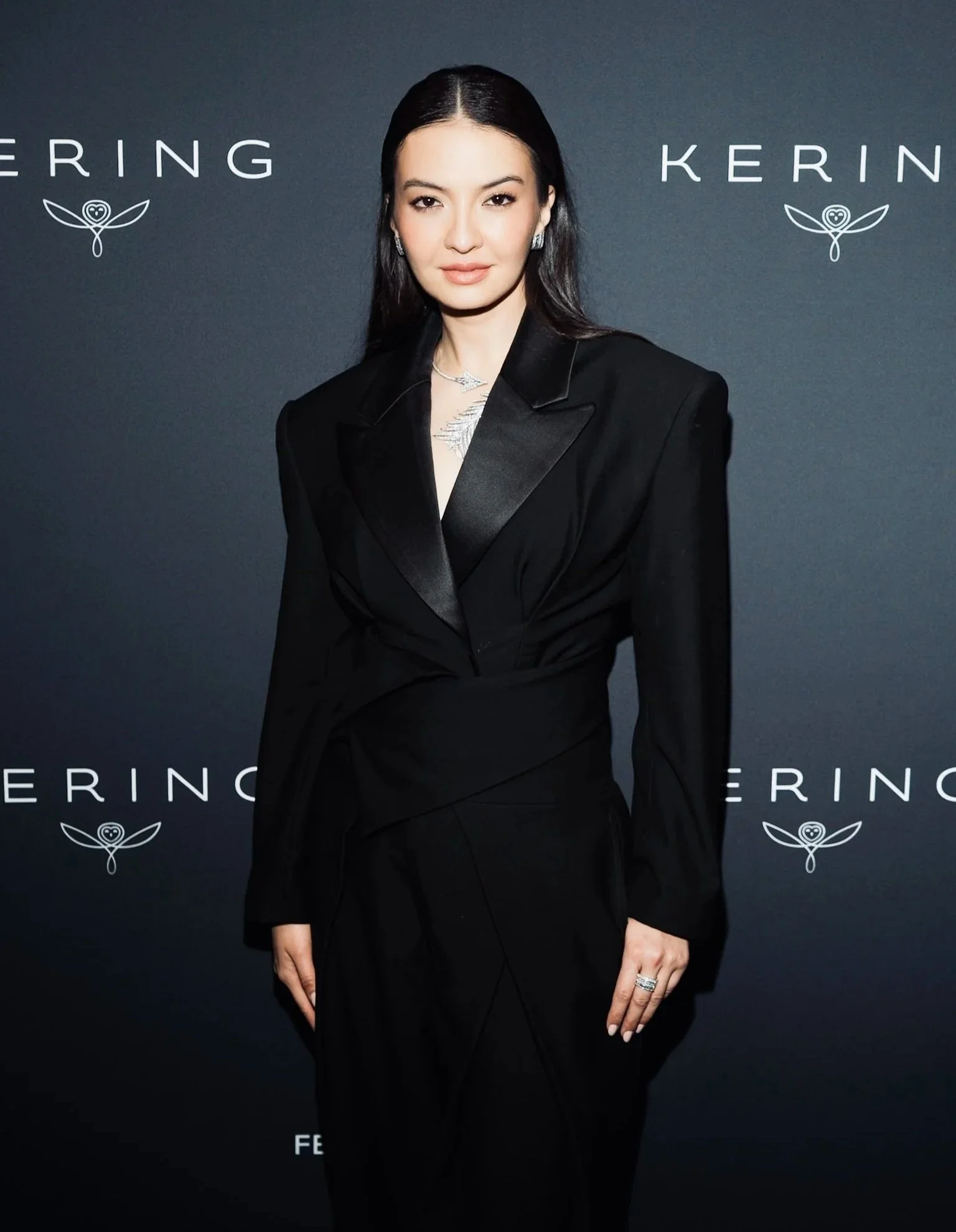 A woman with long black hair, wearing a black blazer and jewelry, standing in front of a gray background with the name 'Kering' and a logo featuring a stylized owl and leaves.