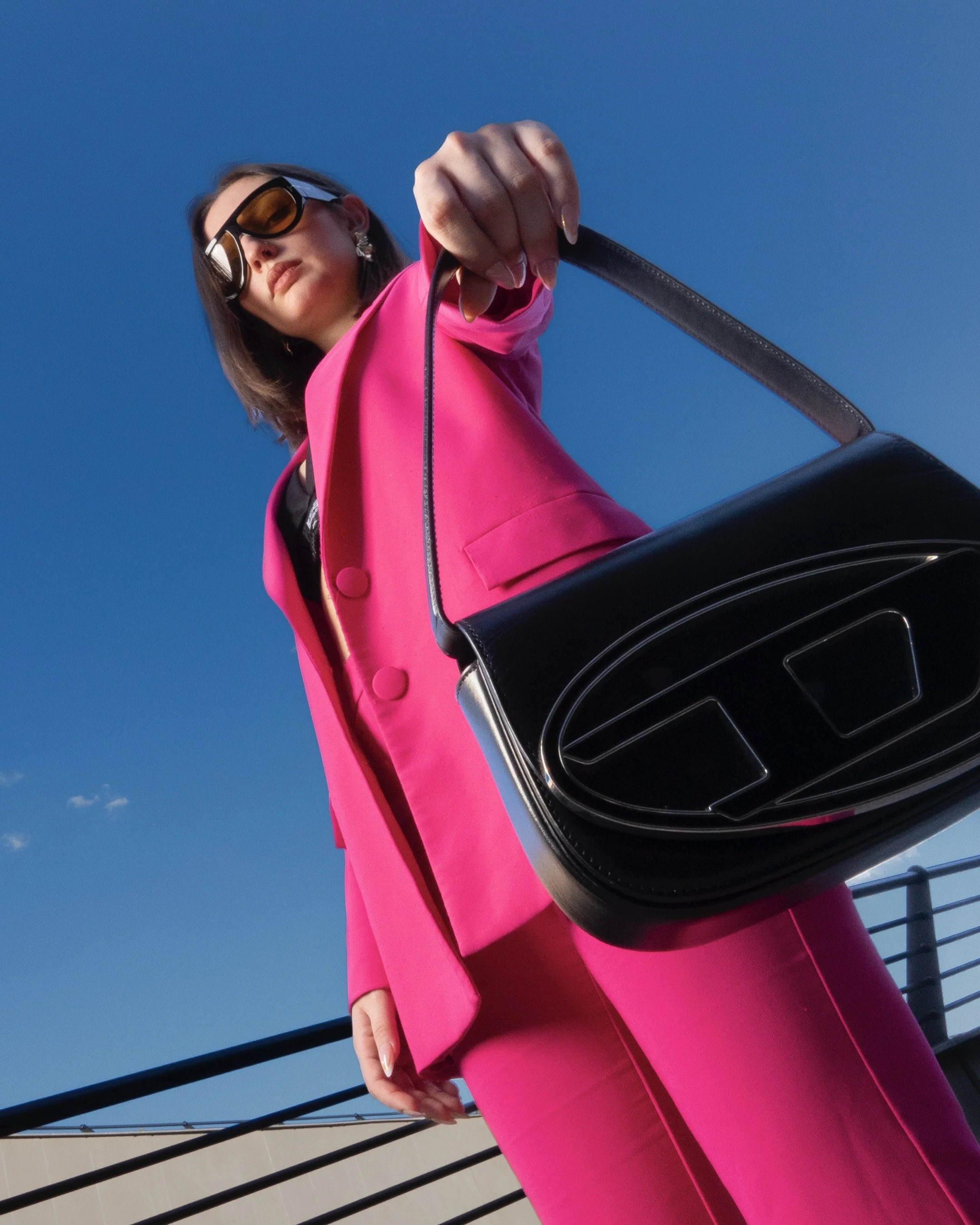 Woman in bright pink suit holding a black Diesel handbag, wearing oversized sunglasses, standing outdoors under a clear blue sky.