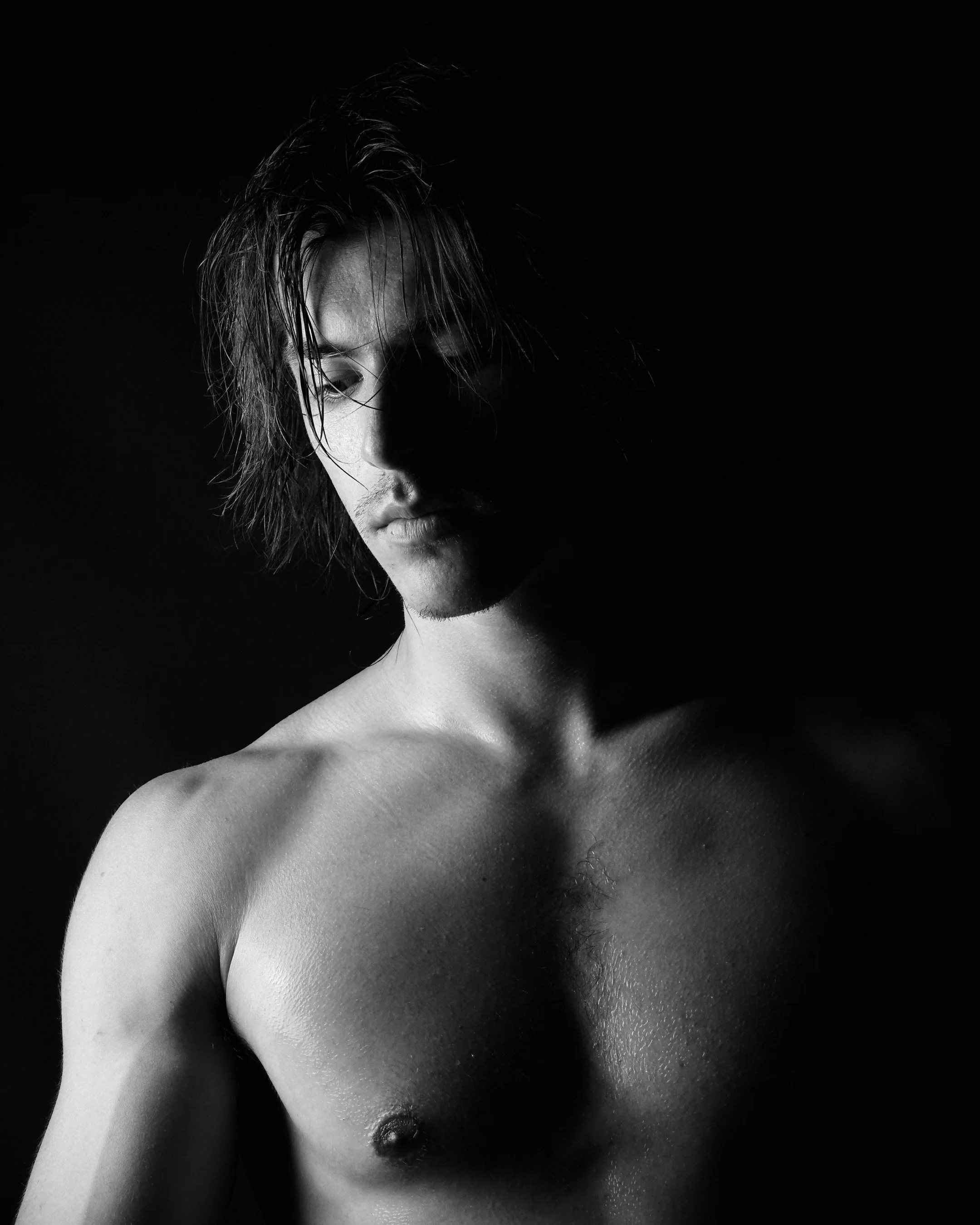 Black and white photo of a Francesco Pividori topless with wet hair, partially shadowed face, looking downward against dark background.