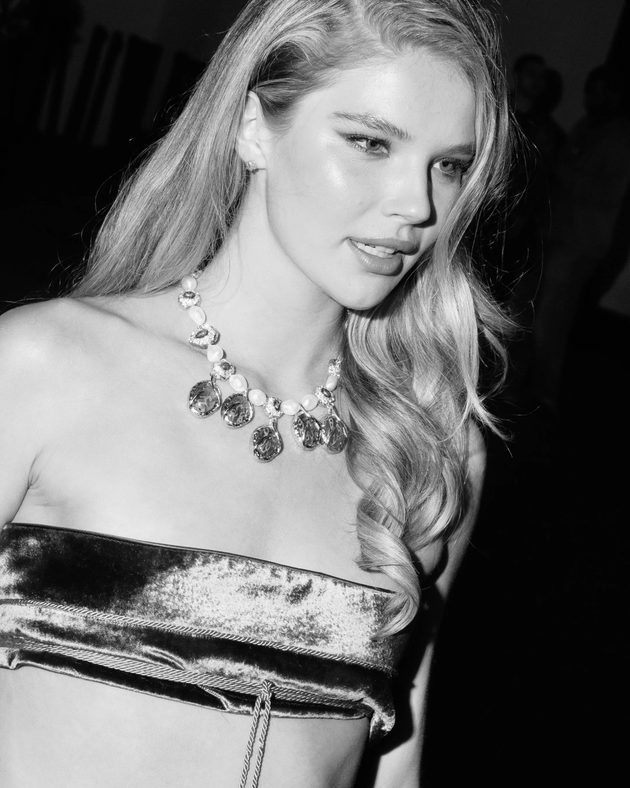 Model Ellie Thumann, a young woman with wavy hair wearing a strapless velvet top and a necklace with large gemstones, smiling slightly with her lips parted.