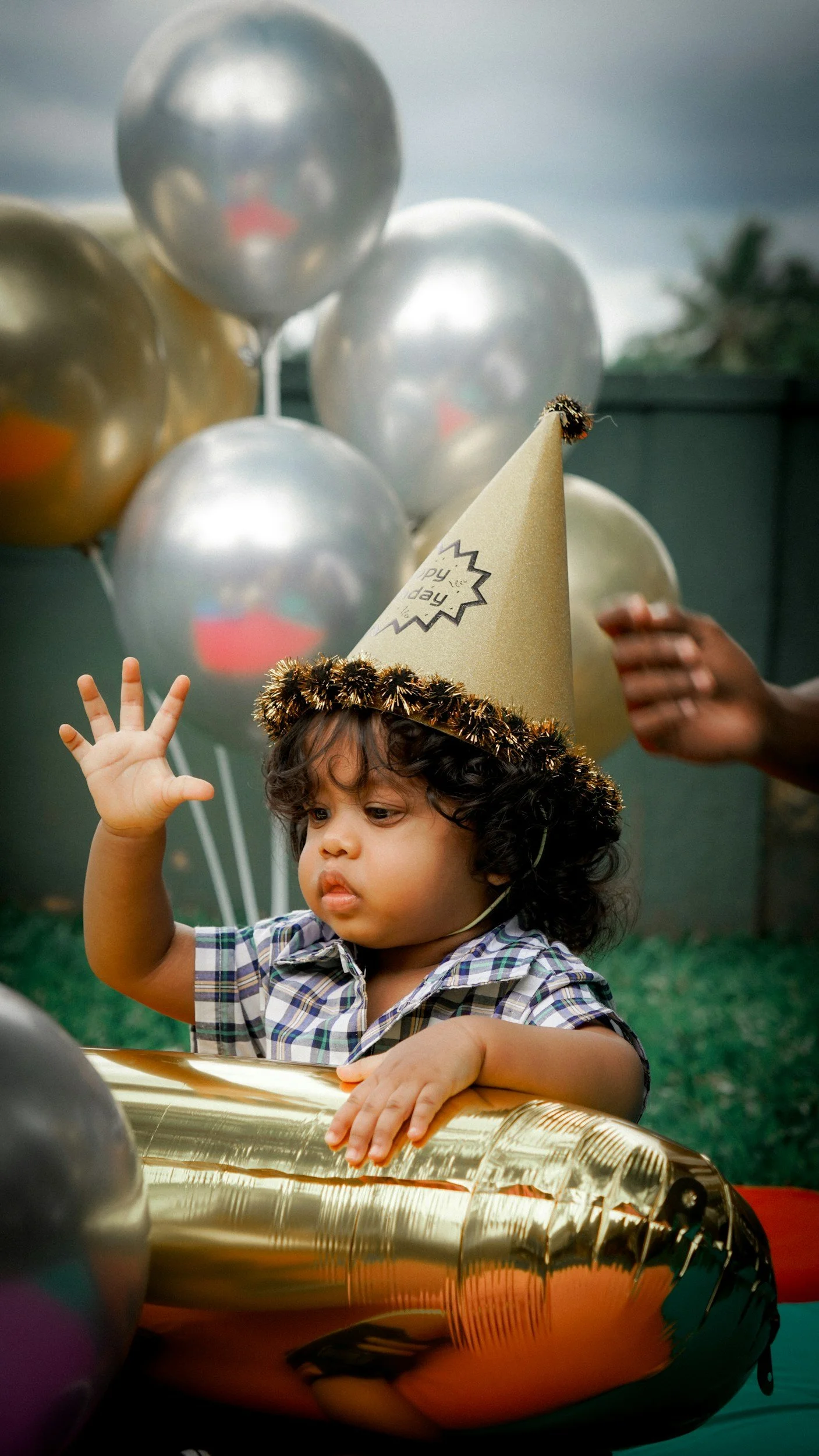 A young child wearing a party hat and plaid shirt sitting in a gold foil balloon boat, surrounded by silver and gold balloons, celebrating a birthday.