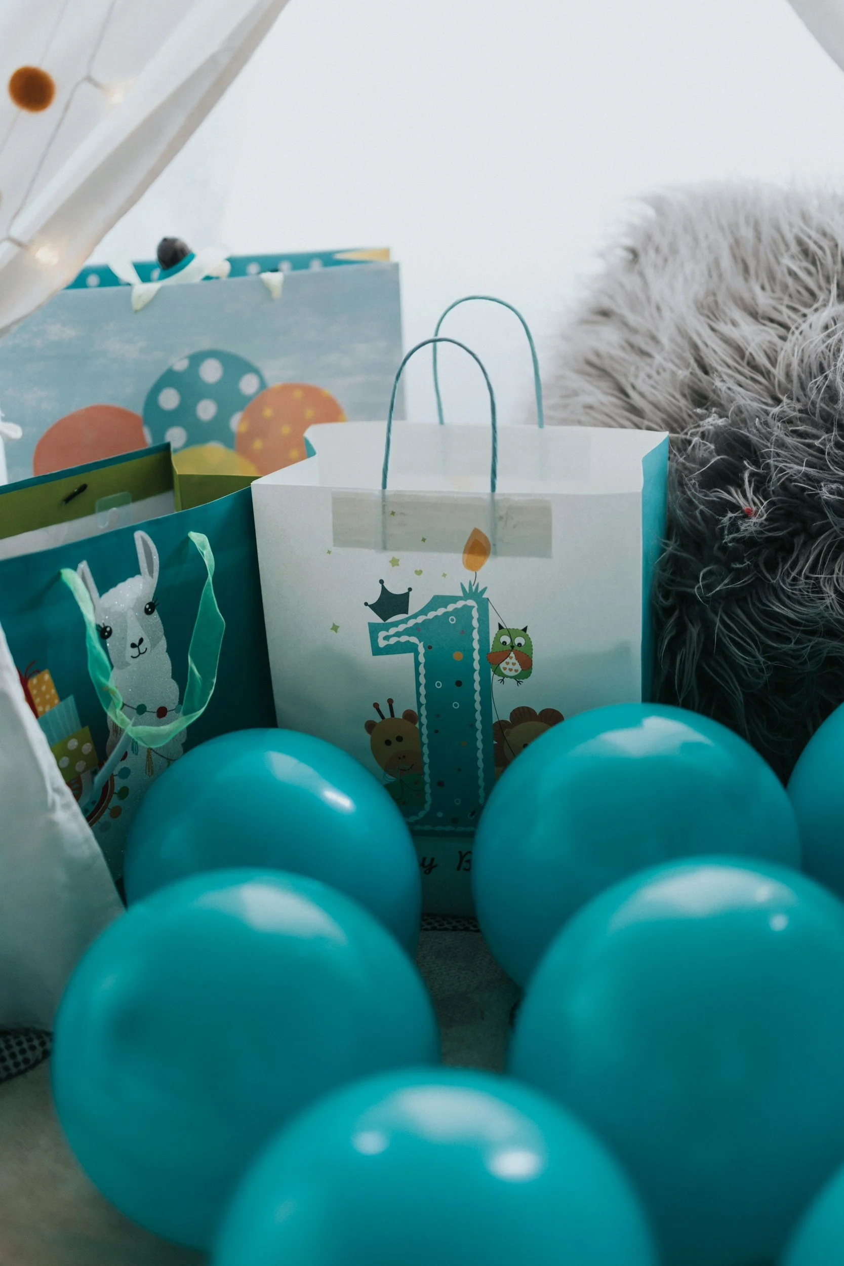 Blue balloons surrounding a birthday gift bag with the number 1 on it and a giraffe, owl, and crown illustration, indicating a first birthday celebration.