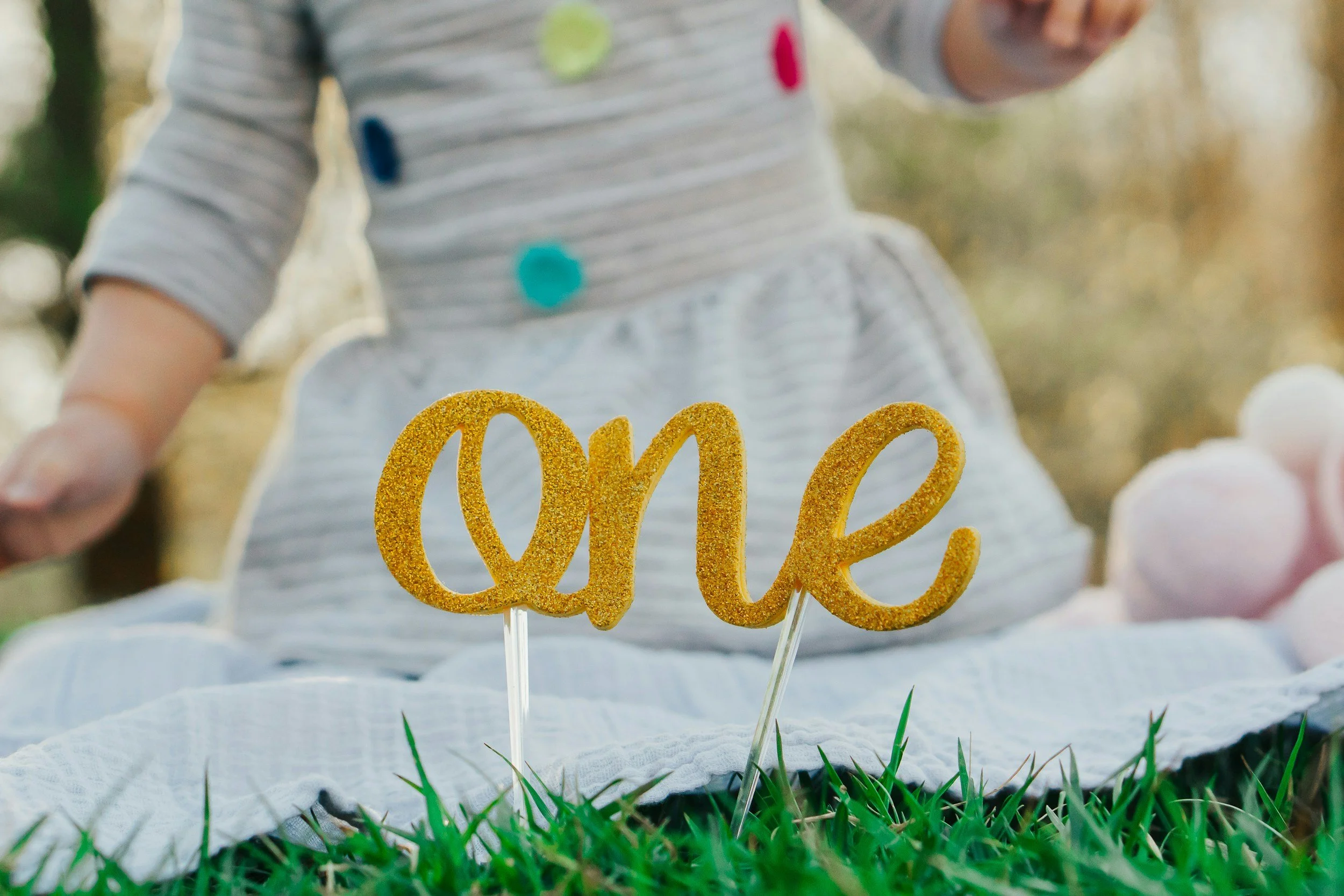 Close-up of a glittery gold 'one' cake topper on a stick, placed on green grass, with a child in a striped dress sitting in the background.