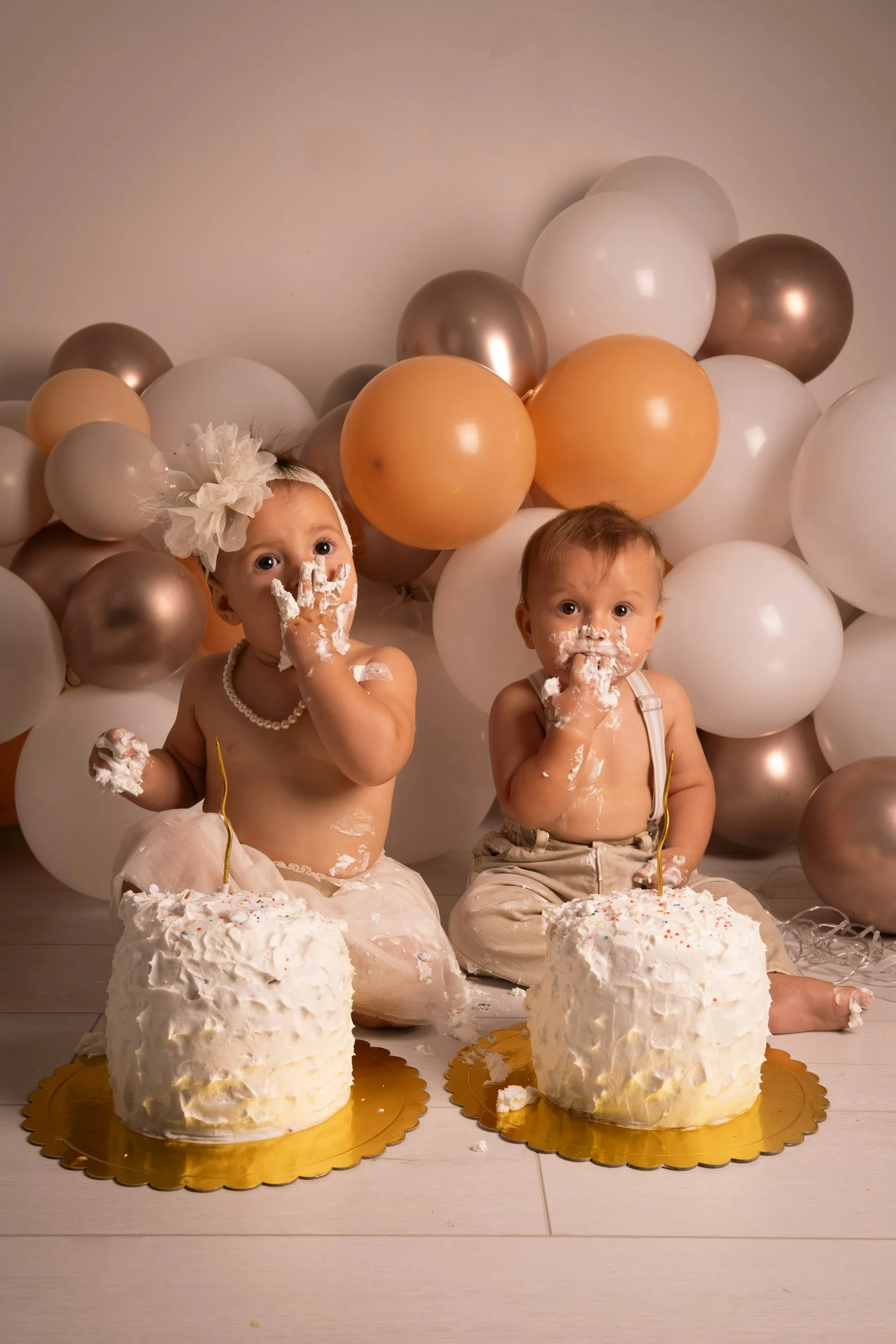 Two babies celebrating a birthday with cakes and balloons in the background.