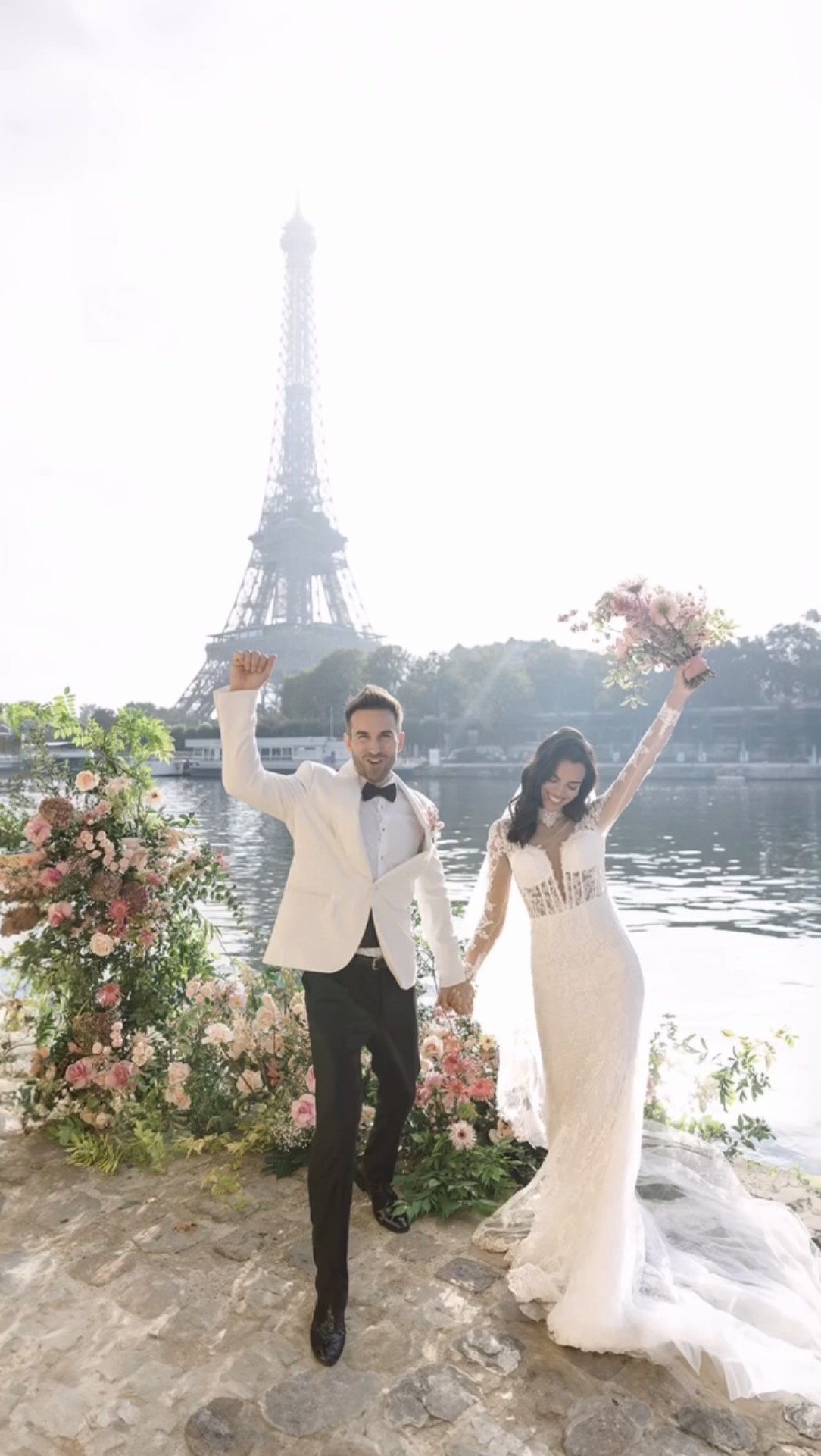 A newlywed couple celebrating by a river with the Eiffel Tower in the background. The groom is wearing a tuxedo, and the bride is in a white lace wedding dress holding a bouquet. They are holding hands and smiling, surrounded by flowers.