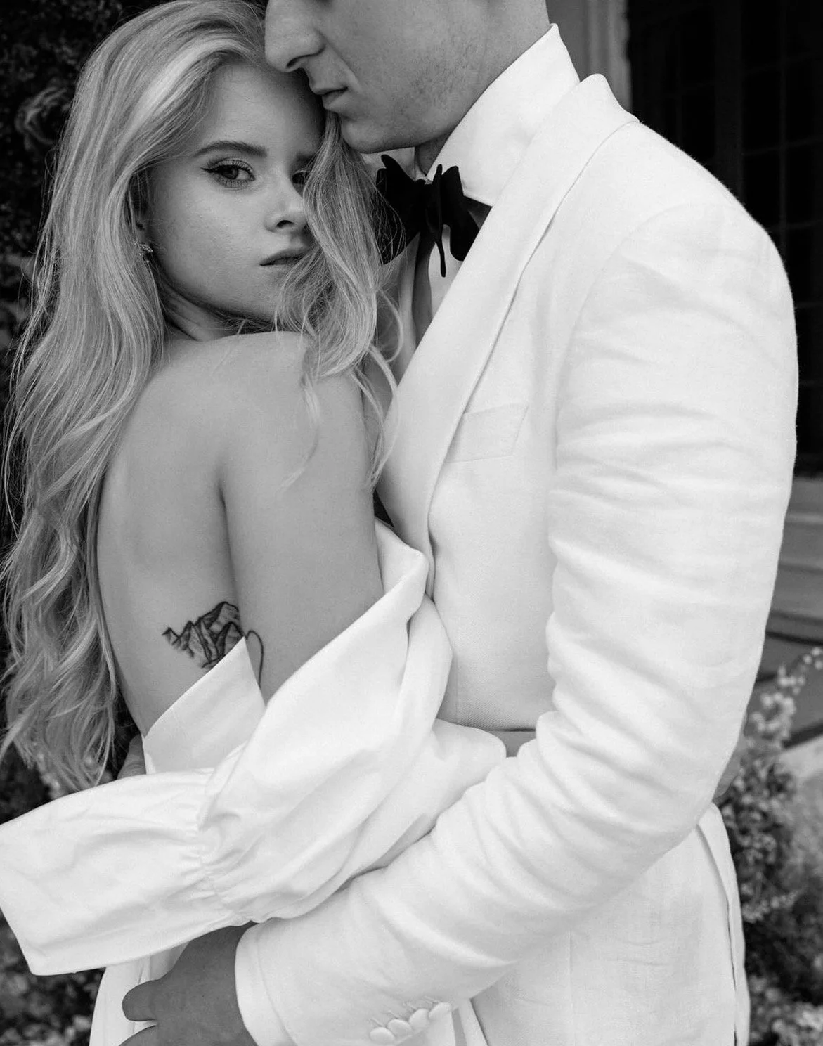 Black and white photo of a man and woman embracing closely, with the man wearing a white tuxedo and the woman in a strapless dress with long wavy hair. The woman has a tattoo on her upper arm.
