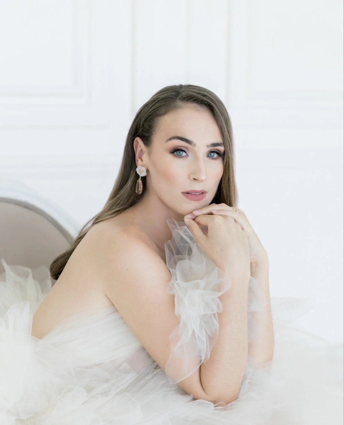 A woman with long brown hair and blue eyes, wearing elegant earrings, sits with her chin resting on her hands, dressed in a white tulle dress, in a bright, white room.