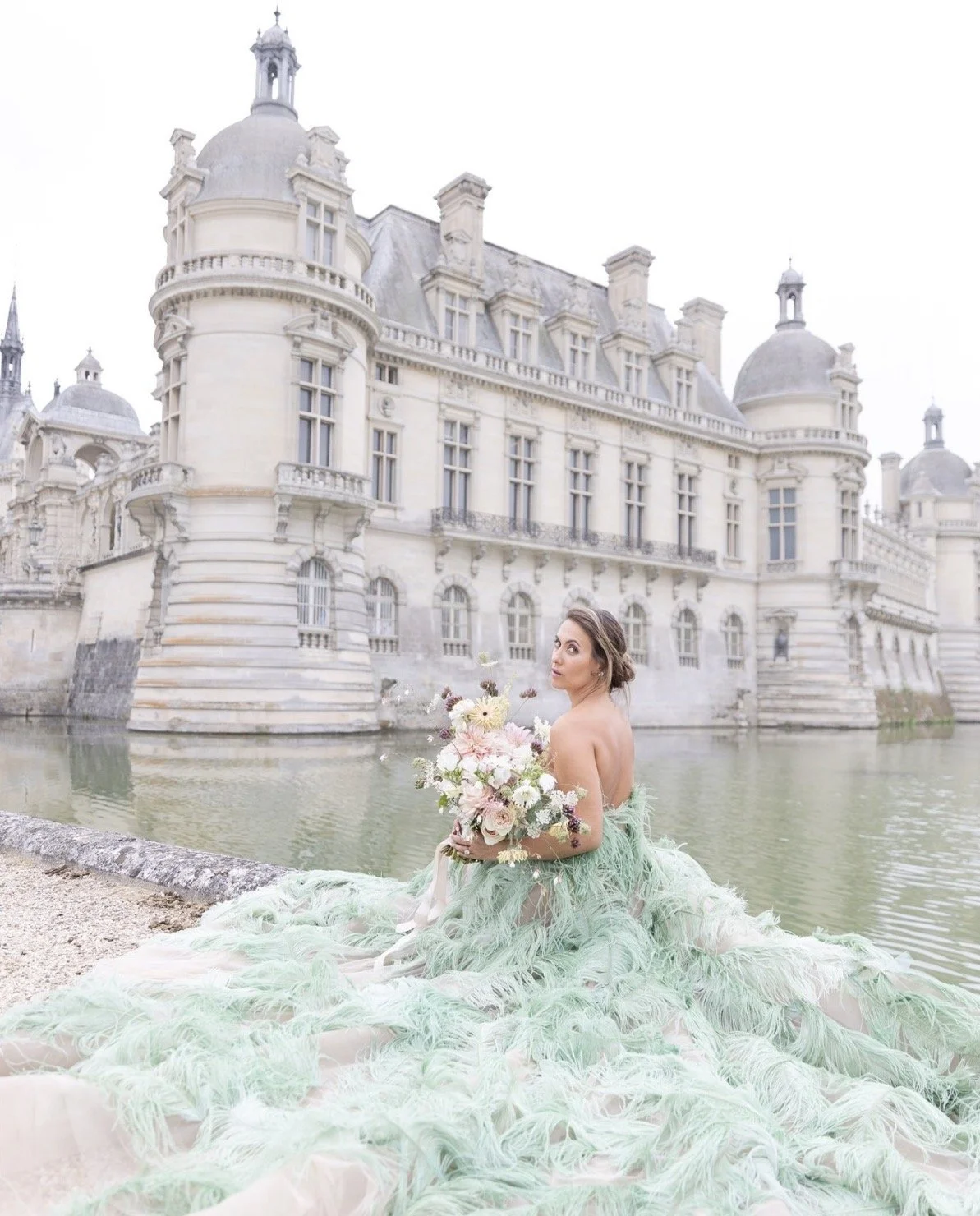 A woman in a green feathered gown holding a bouquet of pink and white flowers, sitting by a moat in front of a grand historic castle.