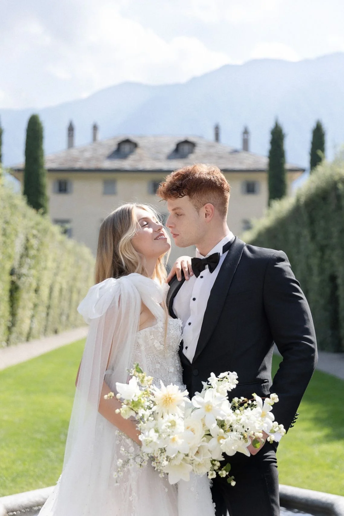 A bride and groom in wedding attire standing outdoors in front of a large house and mountainous background. The bride is holding a bouquet of white flowers and looking up at the groom, who is gazing down at her. EUROPE WEDDING