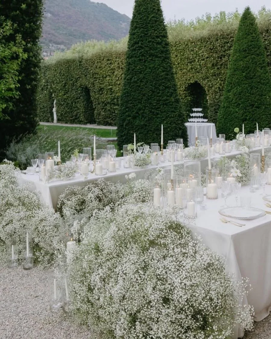 Elegant outdoor dinner setup with white tablecloths, tall white candles, and floral arrangements, set in a garden with tall hedges and manicured trees.