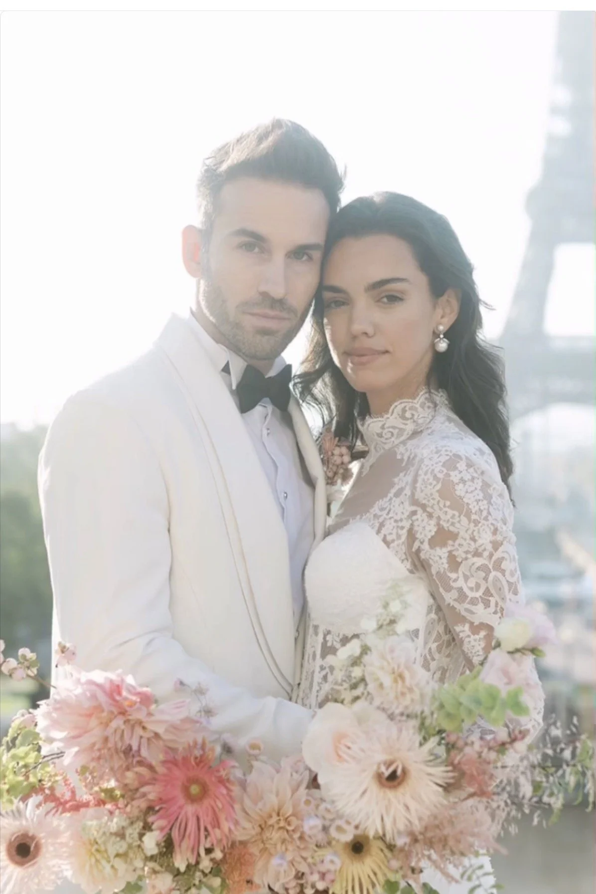A couple dressed in wedding attire, standing close together outdoors, with a large bouquet of pink, white, and peach flowers in the foreground. The background shows a cityscape with the Eiffel Tower.