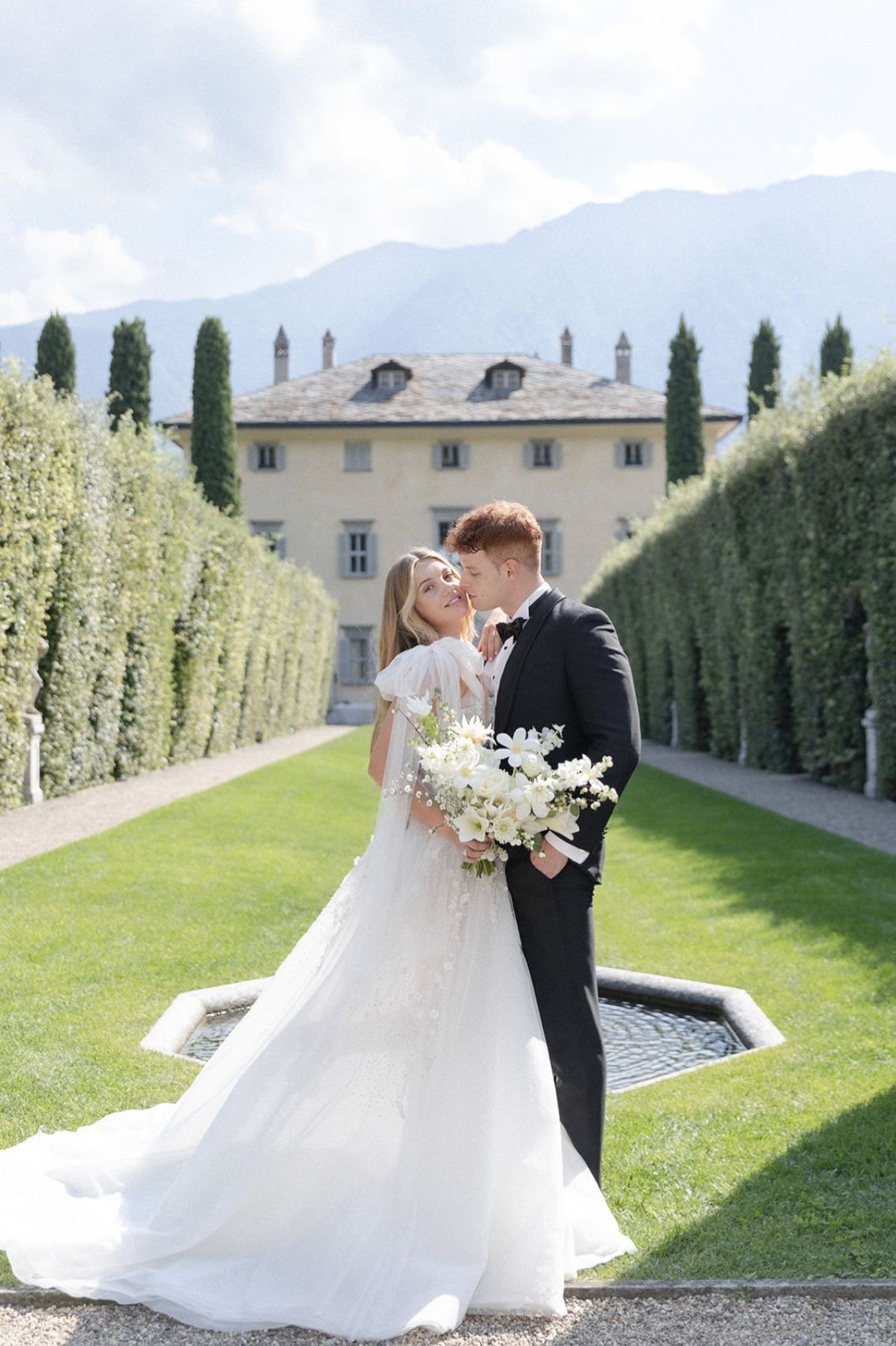 A bride and groom in wedding attire sharing a kiss outdoors on a green lawn with a historic building and mountain backdrop.