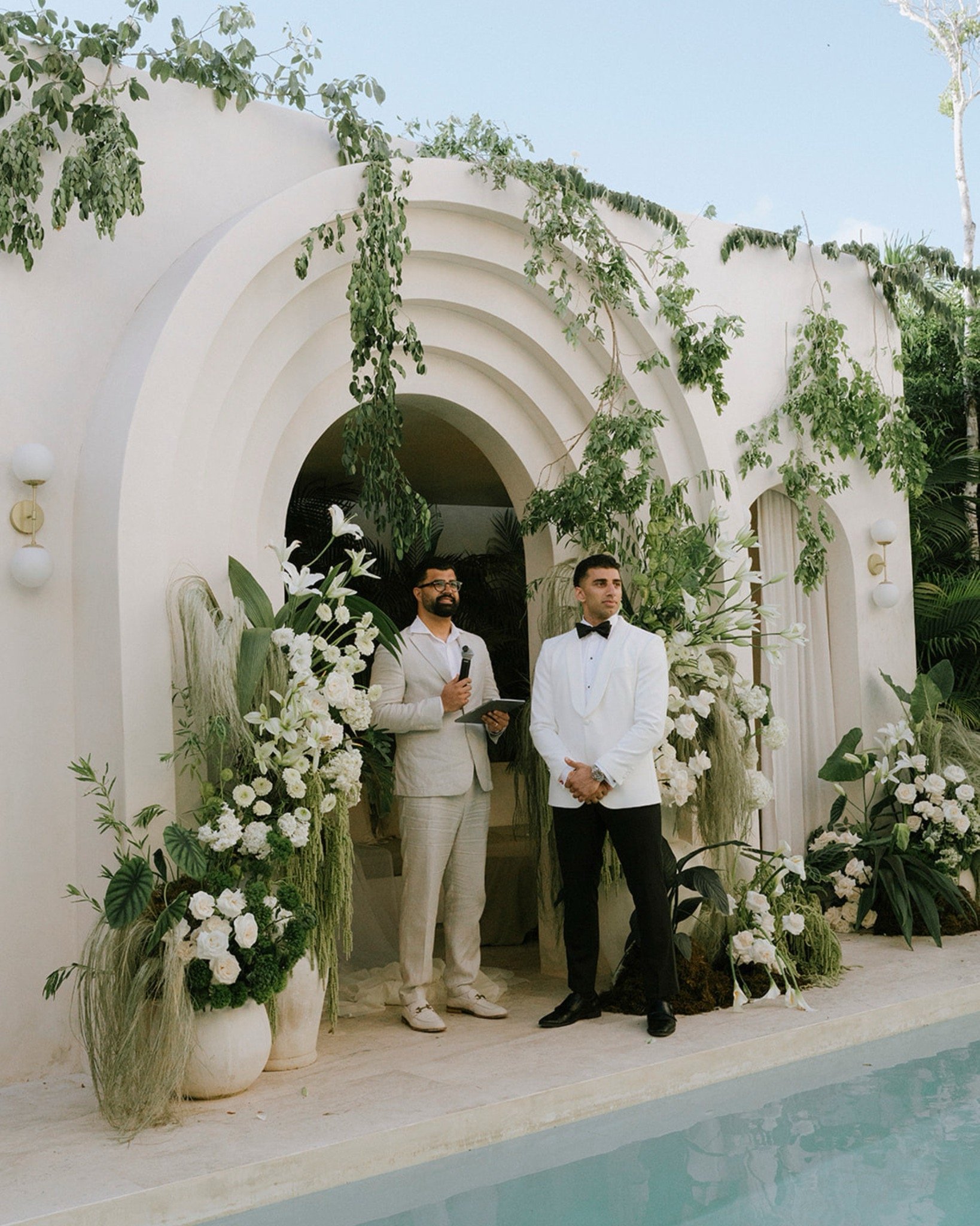 Two men dressed in formal attire standing in an elegant outdoor setting decorated with abundant white flowers and green foliage, next to white urn planters and a pool.