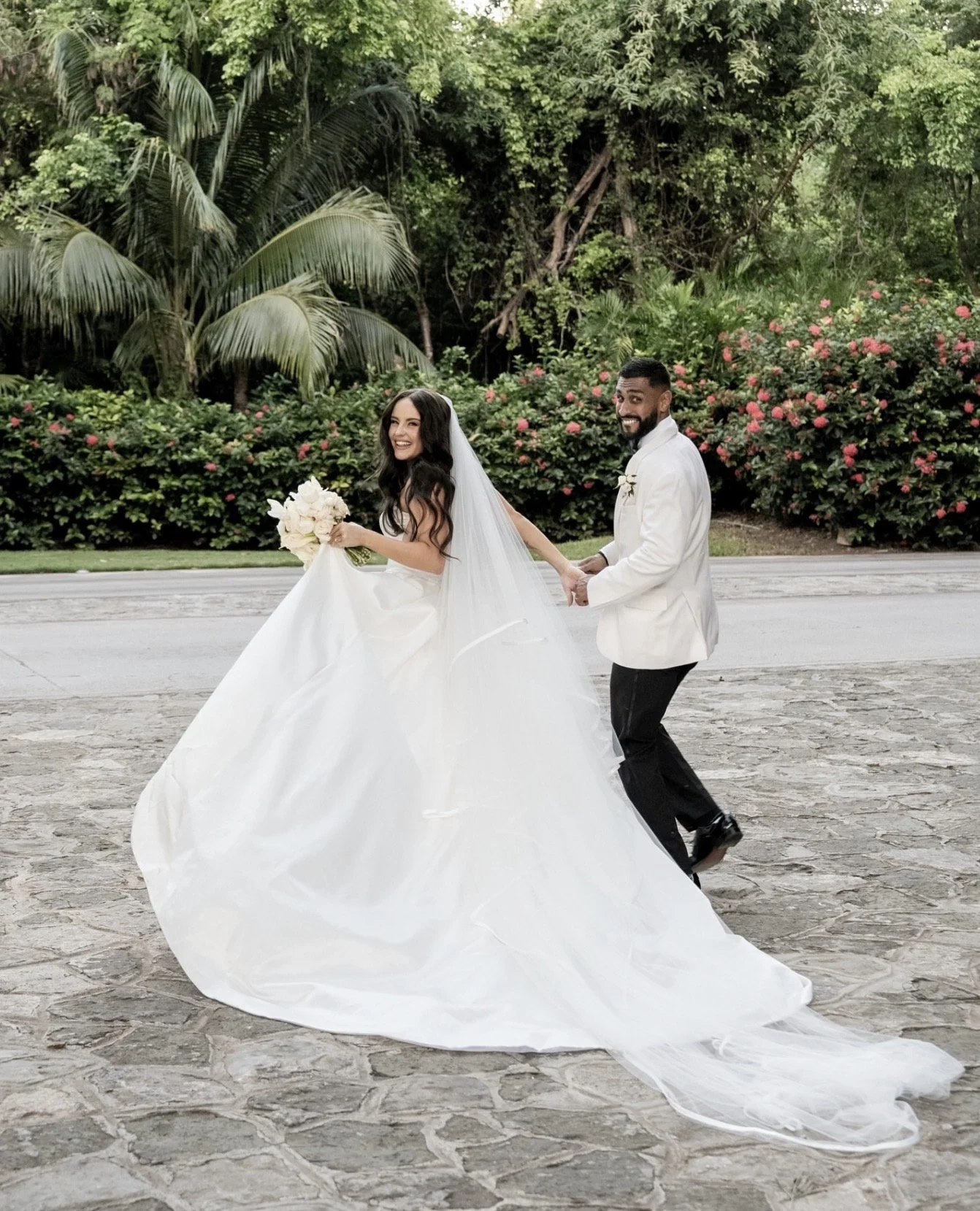 A bride and groom holding hands, smiling, outdoors on a stone paved area with lush greenery and flowers in the background.