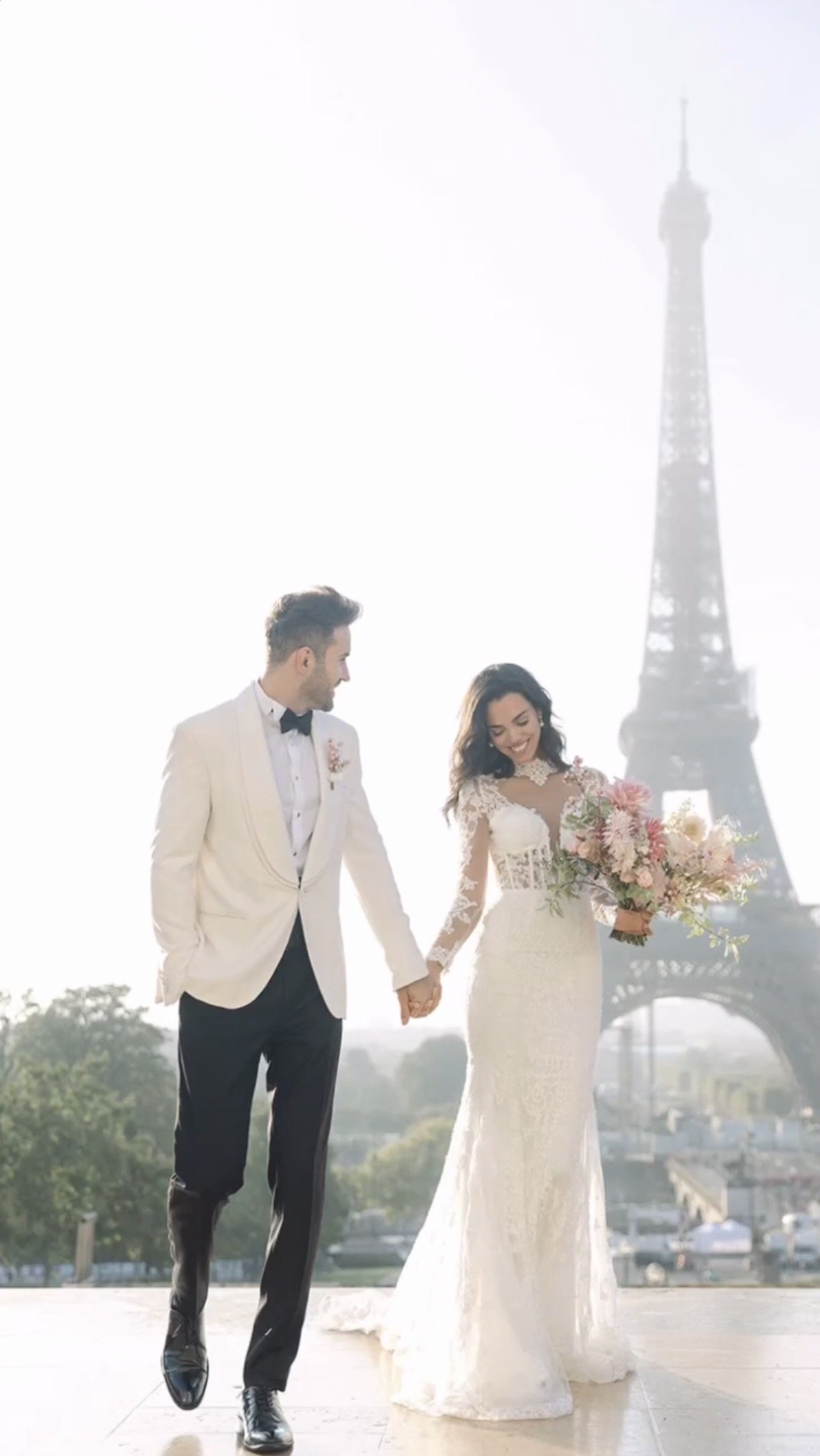 A newlywed couple holding hands and walking near the Eiffel Tower in Paris, France. The bride is in a white lace wedding gown holding a bouquet of pink and white flowers. The groom is in a white tuxedo jacket with black pants and a bow tie.