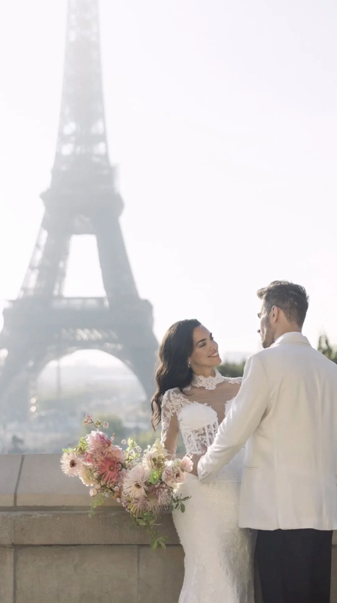 A bride and groom in wedding attire holding hands with the Eiffel Tower in the background.