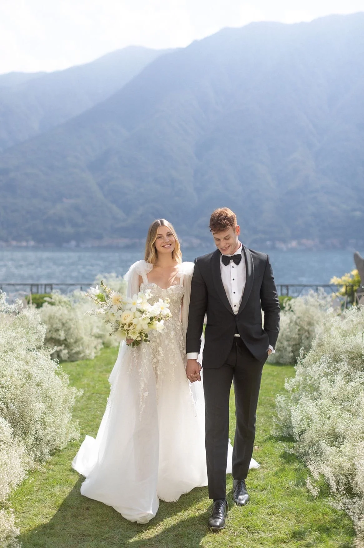 A bride and groom holding hands and walking outdoors near a lake, with mountains in the background, during a wedding ceremony. The bride is wearing a white wedding dress with lace details, and the groom is dressed in a black tuxedo with a bow tie.