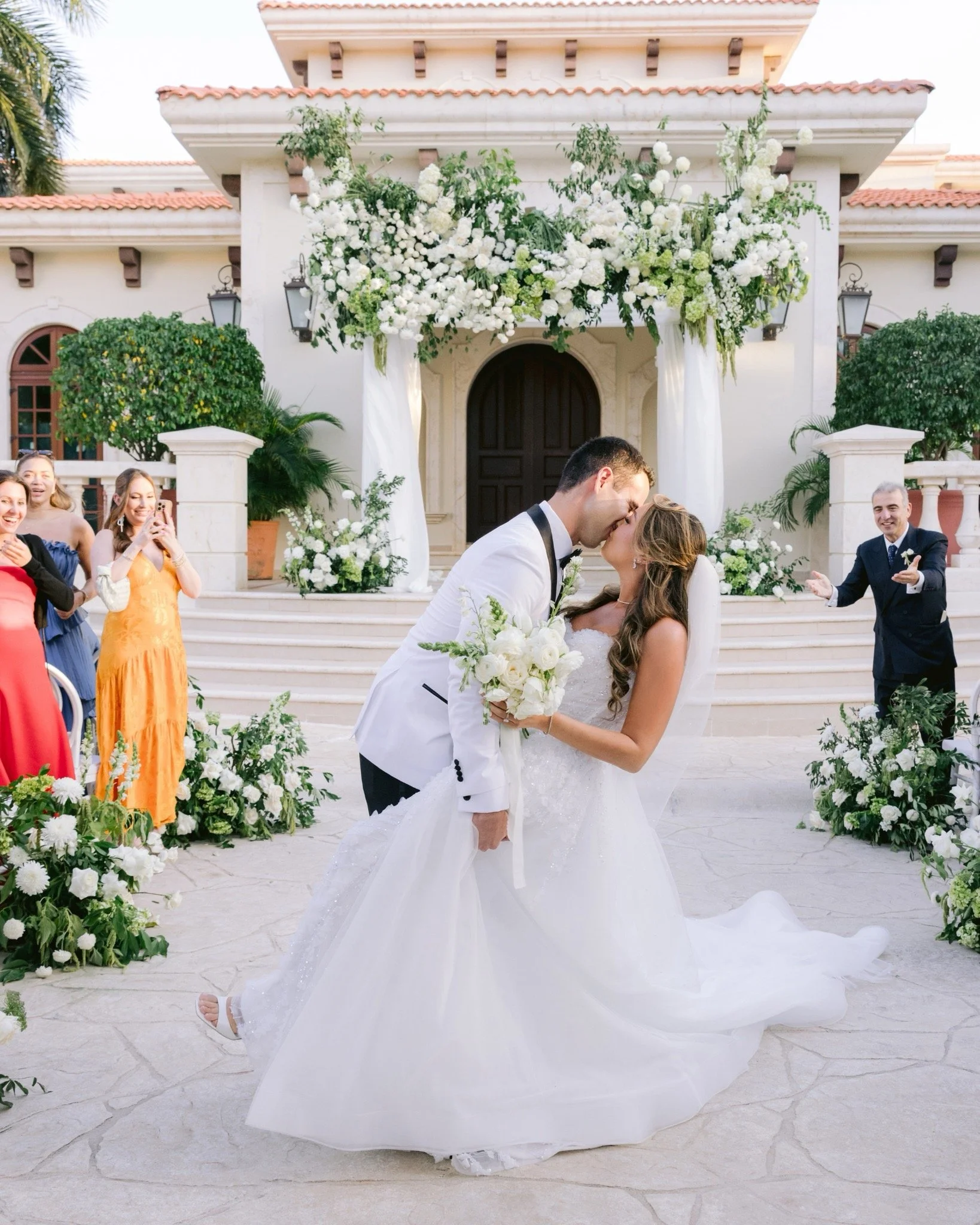 A bride and groom kiss during their wedding ceremony outdoors, decorated with white flowers and greenery, in front of a tiled-roof building with white walls and wooden accents.