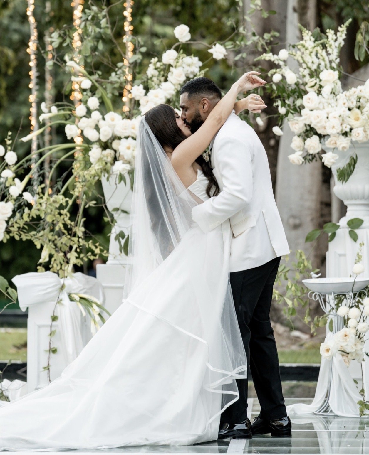 A bride and groom kissing during their outdoor wedding, surrounded by white flowers and greenery.