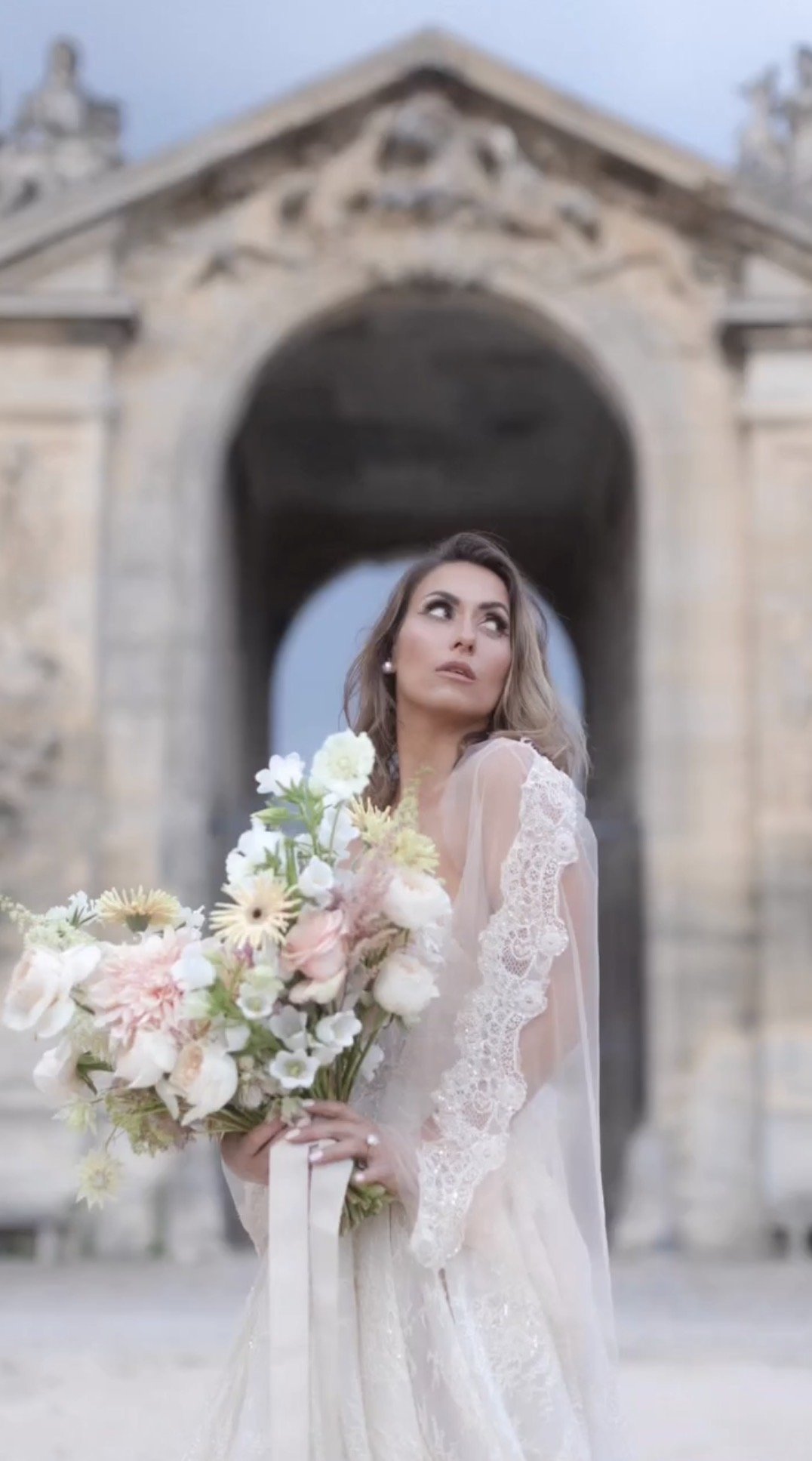 A woman in a wedding dress holding a bouquet of flowers in front of a historic stone archway.