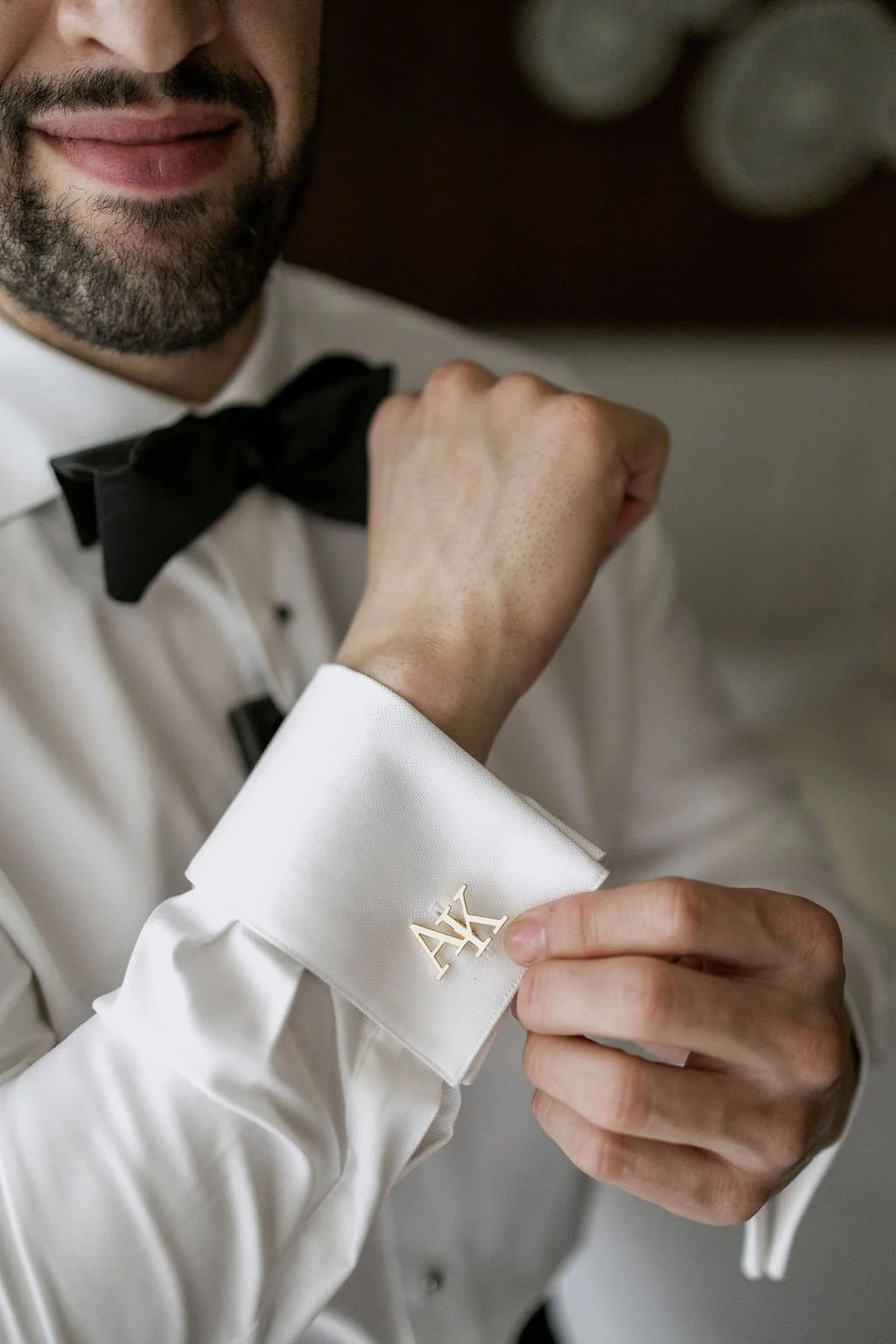 A man with a beard and mustache, dressed in a white tuxedo with a black bow tie, adjusting his cuff with a gold cufflink engraved with the letters 'A' and 'K'.