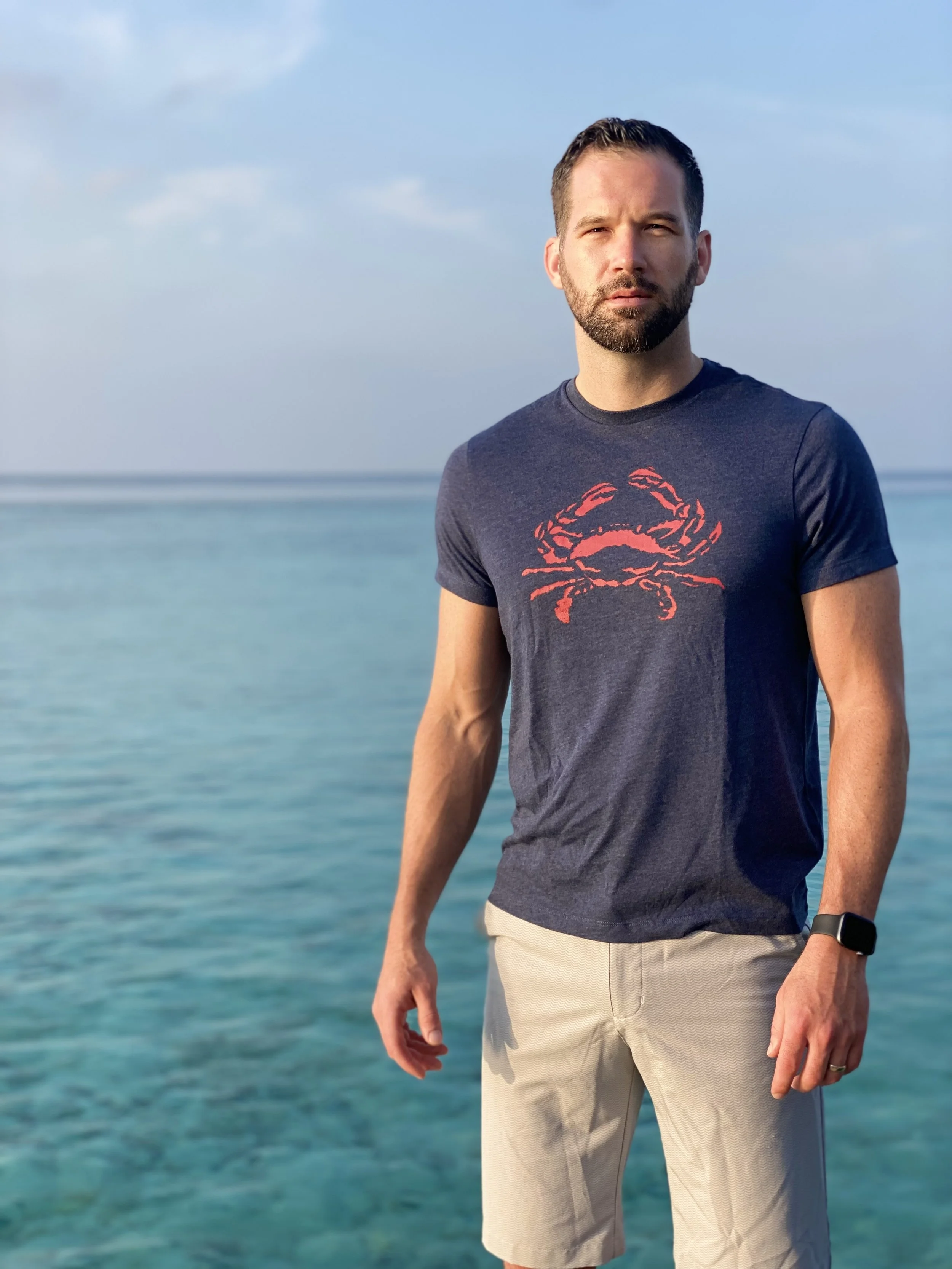 Man with beard in navy blue t-shirt with crab graphic, beige shorts, standing by the ocean with blue sky and clouds. Maldives
