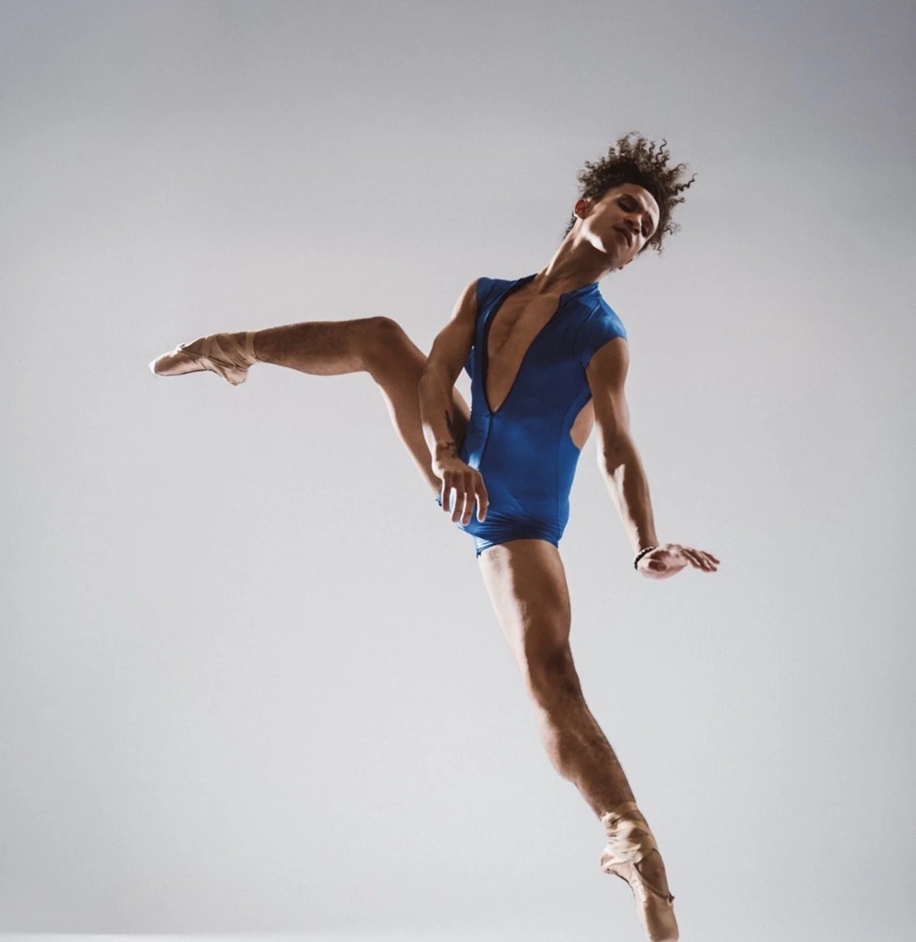 A trans female ballet dancer in a blue costume performing a dance move against a plain background.