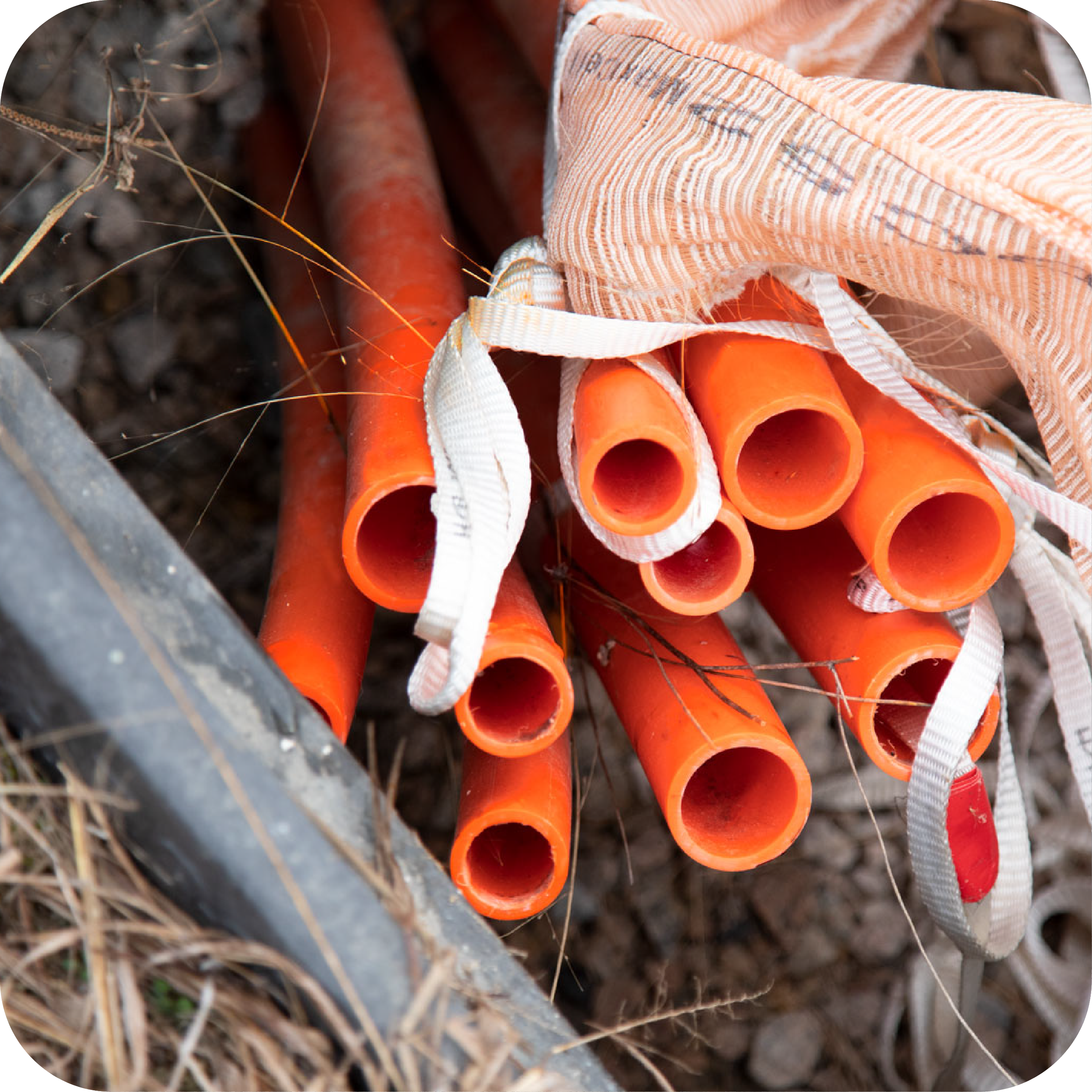 A bundle of orange conduit pipes tied with white and red straps, lying on dirt with some dry grass and a metal object nearby.