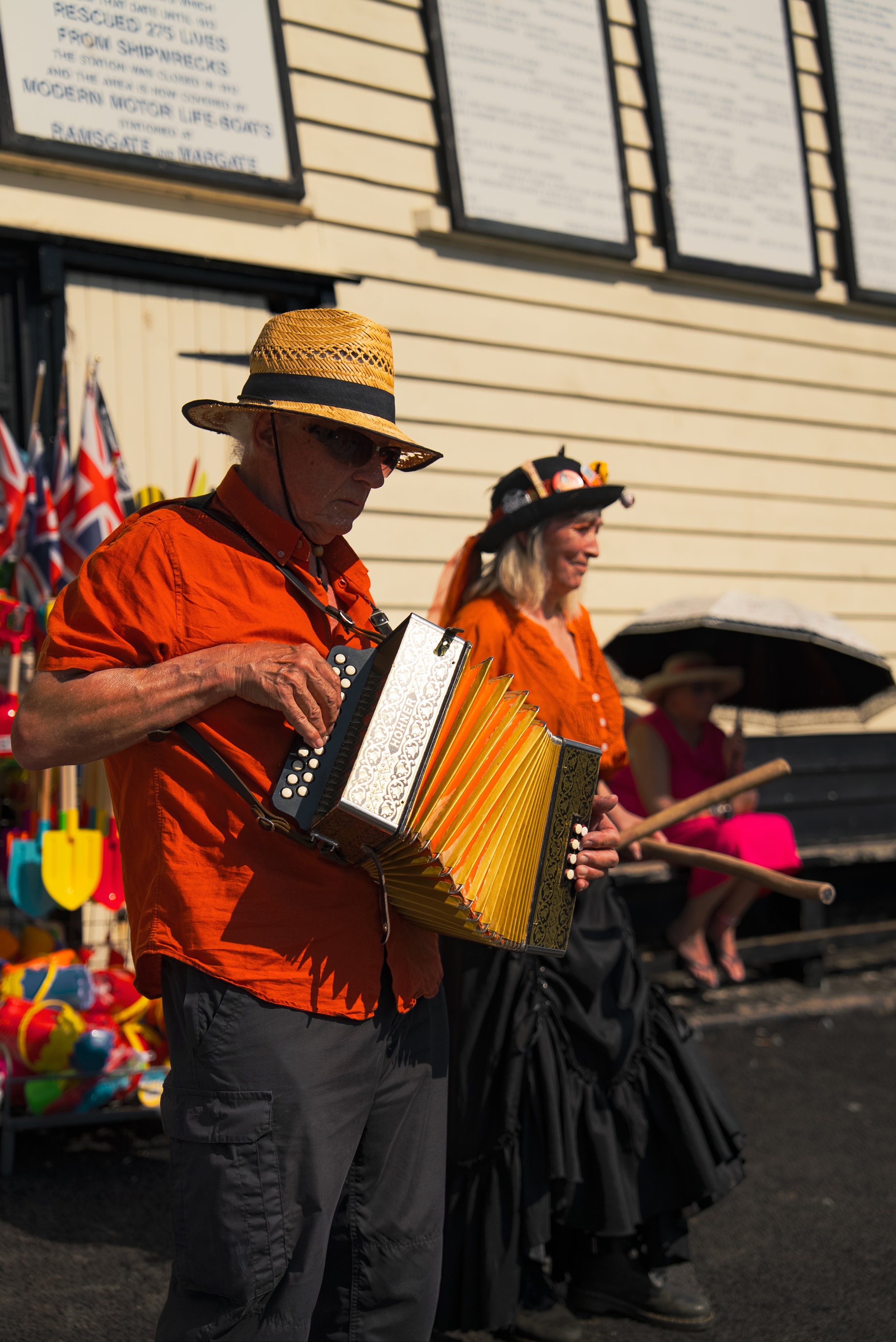 An older man playing an accordion with a woman dressed in black behind him, both dressed in orange shirts; the man wears a straw hat and glasses, the woman wears a black hat with pins. They are outdoors near a building with signs and small Union Jack