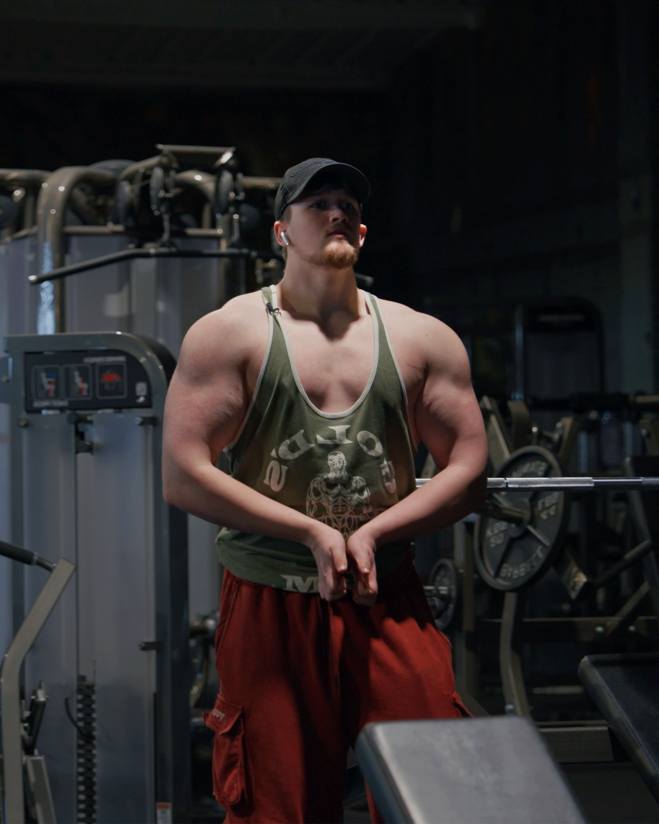 A muscular man in a gym, wearing a black cap, green tank top, and red shorts, standing near gym equipment in a dimly lit setting.