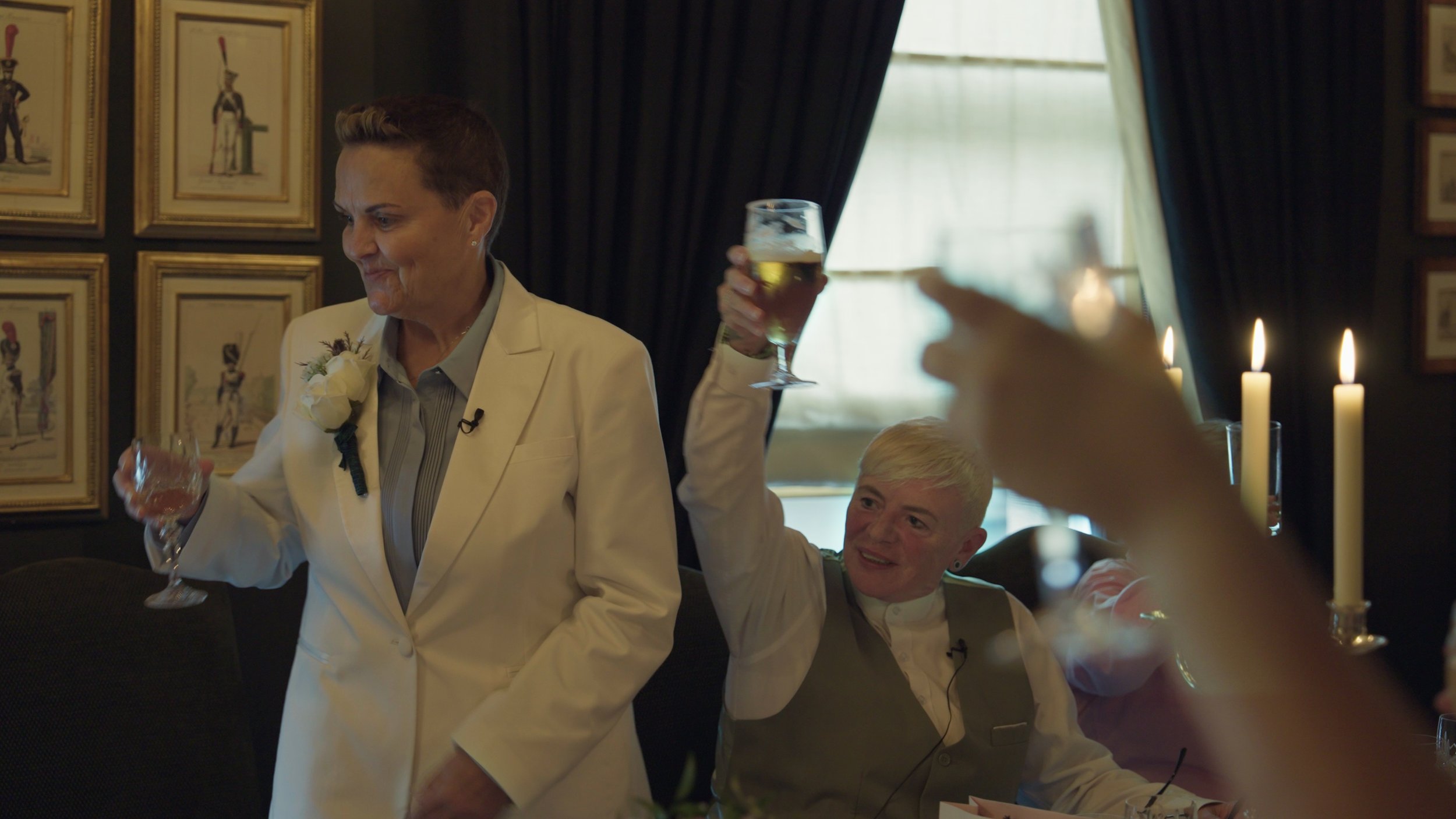 Two women at a celebration, one standing in a white suit with a flower pinned to her lapel, the other sitting and raising a glass of white wine in a toast. The second woman has short blonde hair and is smiling. The setting appears to be a decorated i