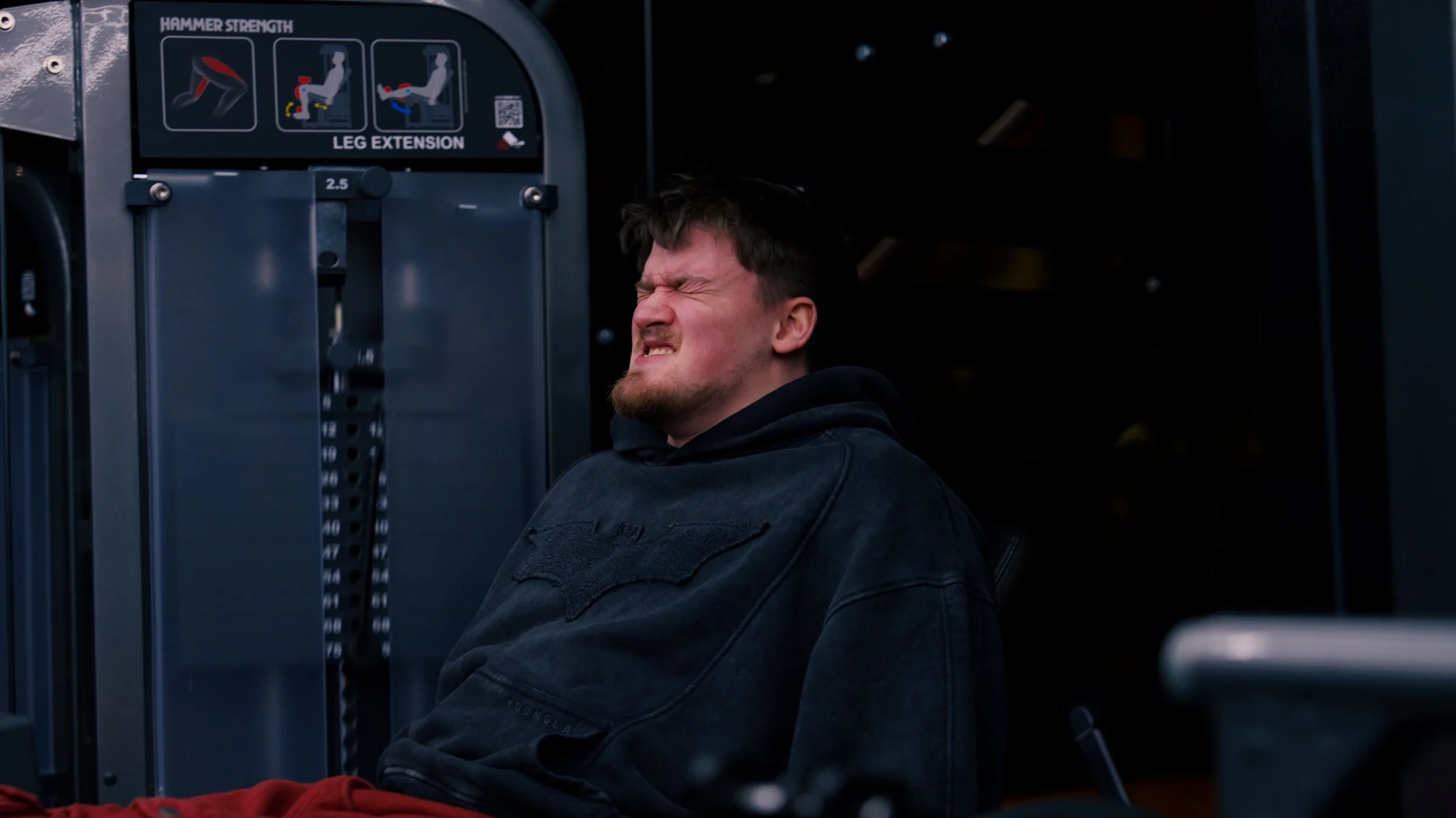 Man grimacing with eyes closed, sitting next to a leg extension workout machine in a gym.